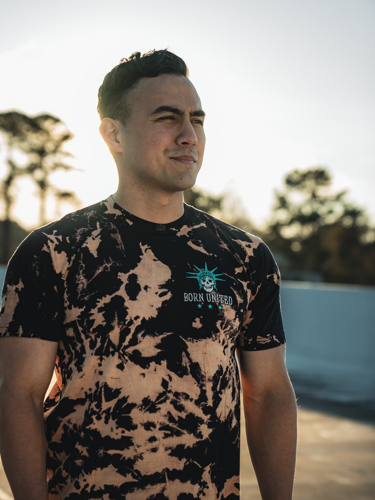 A person stands outside in a Born United Private Label Liberty or Death - Bleach Wash shirt with BORN UN1TED. As the sun sets, casting a warm glow, the spirit of freedom is palpable. Tall trees witness this serene moment from afar.