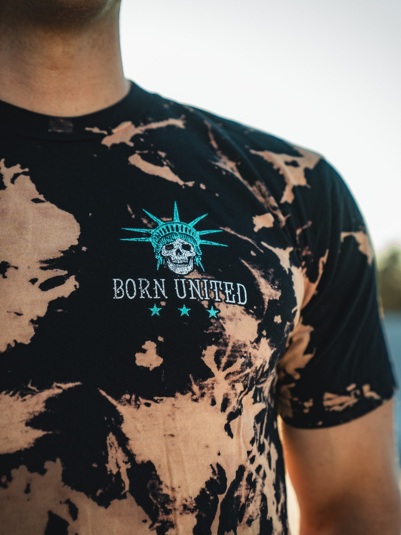 A person wearing the Liberty or Death - Bleach Wash shirt from Born United Private Label. The black and brown tie-dye features a skeleton with a spiked crown above BORN UNITED and three stars, embodying freedom against a light sky backdrop.