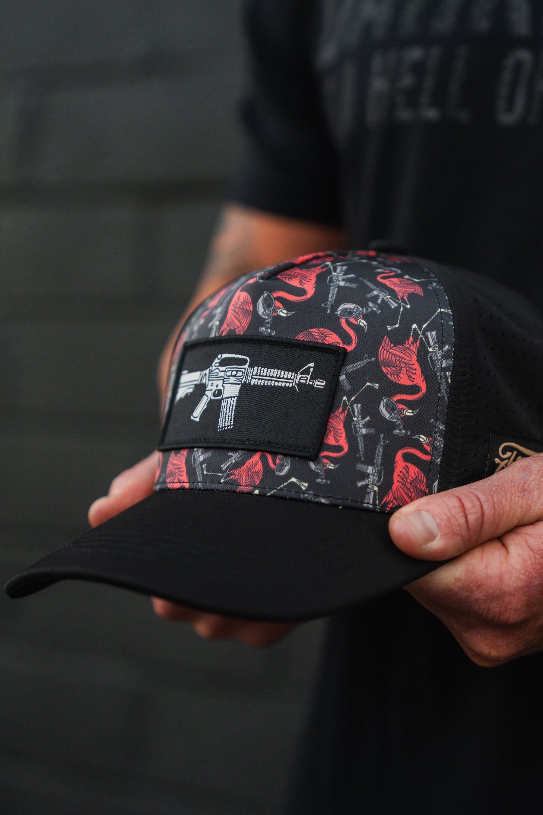 A person holds the Tactimingo Hydro Snapback Hat by Tango Charlie Apparel, featuring a black design with red flamingos and guns. It has a rectangular patch displaying a rifle and is sweat-resistant, ideal for adventures. The blurred background reveals an indistinct wall, enhancing its rugged appeal.