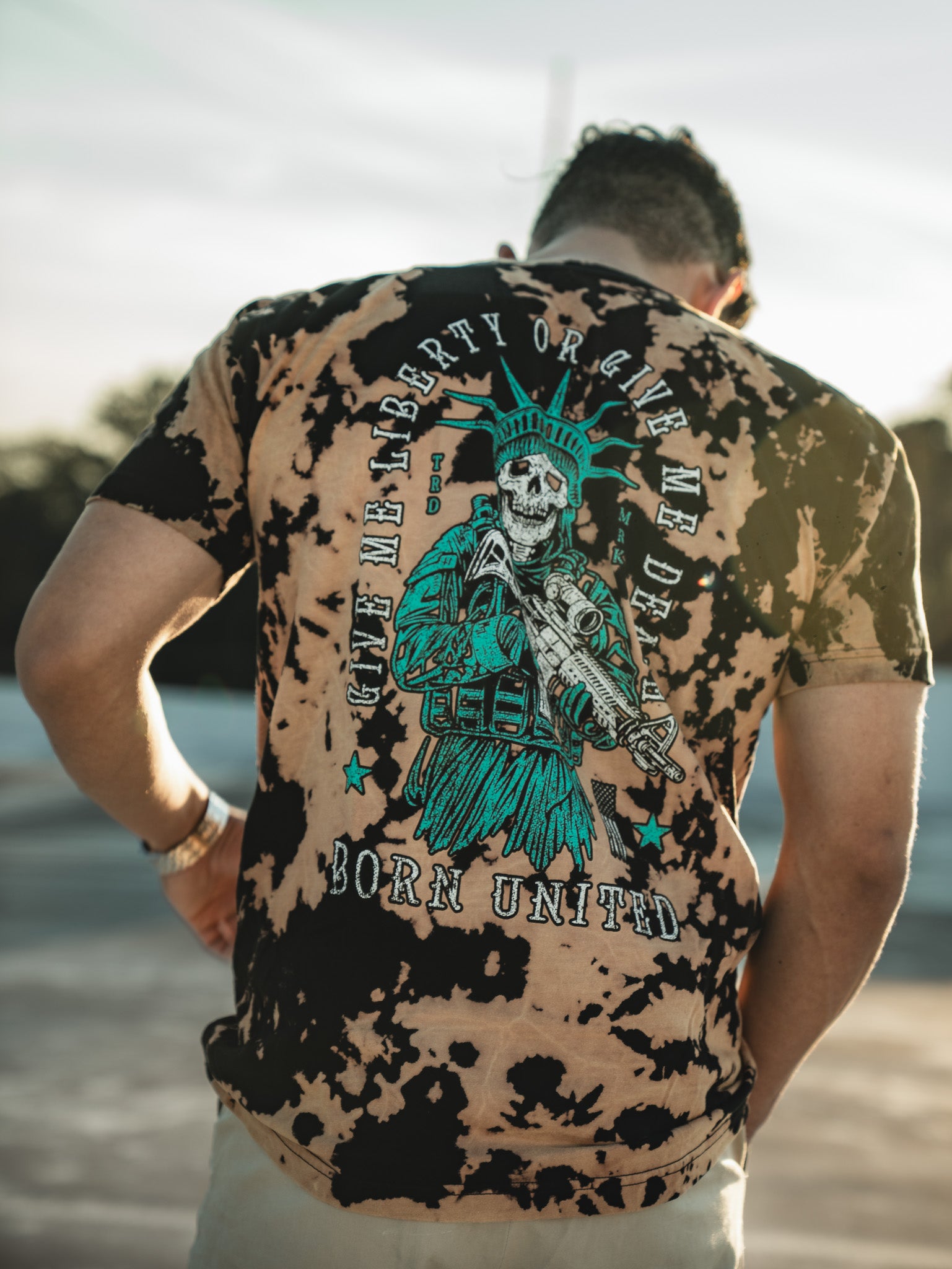 A man wears a Liberty or Death - Bleach Wash T-shirt by Born United Private Label, showcasing a skeleton as the Statue of Liberty with a rifle. The shirt reads Give Me Liberty or Give Me Death and Born United, capturing the freedom-loving spirit in an outdoor setting with a blurred sky.