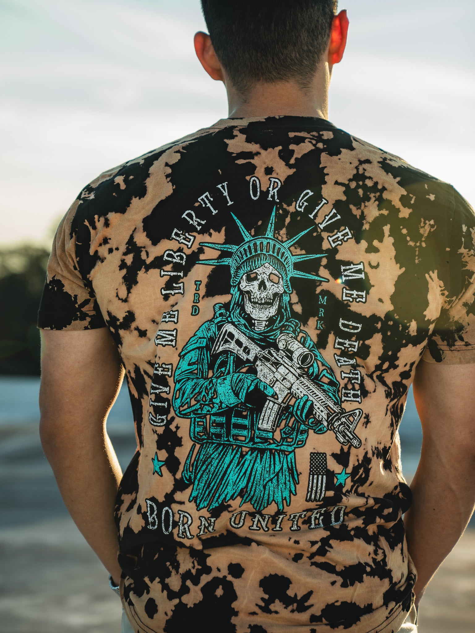 A man wearing the Liberty or Death - Bleach Wash shirt by Born United Private Label. This tie-dye shirt features a back graphic of a skeleton in a Statue of Liberty pose holding a weapon, with the text Give me liberty or give me death, born unto you. A bold statement for patriots and freedom lovers.