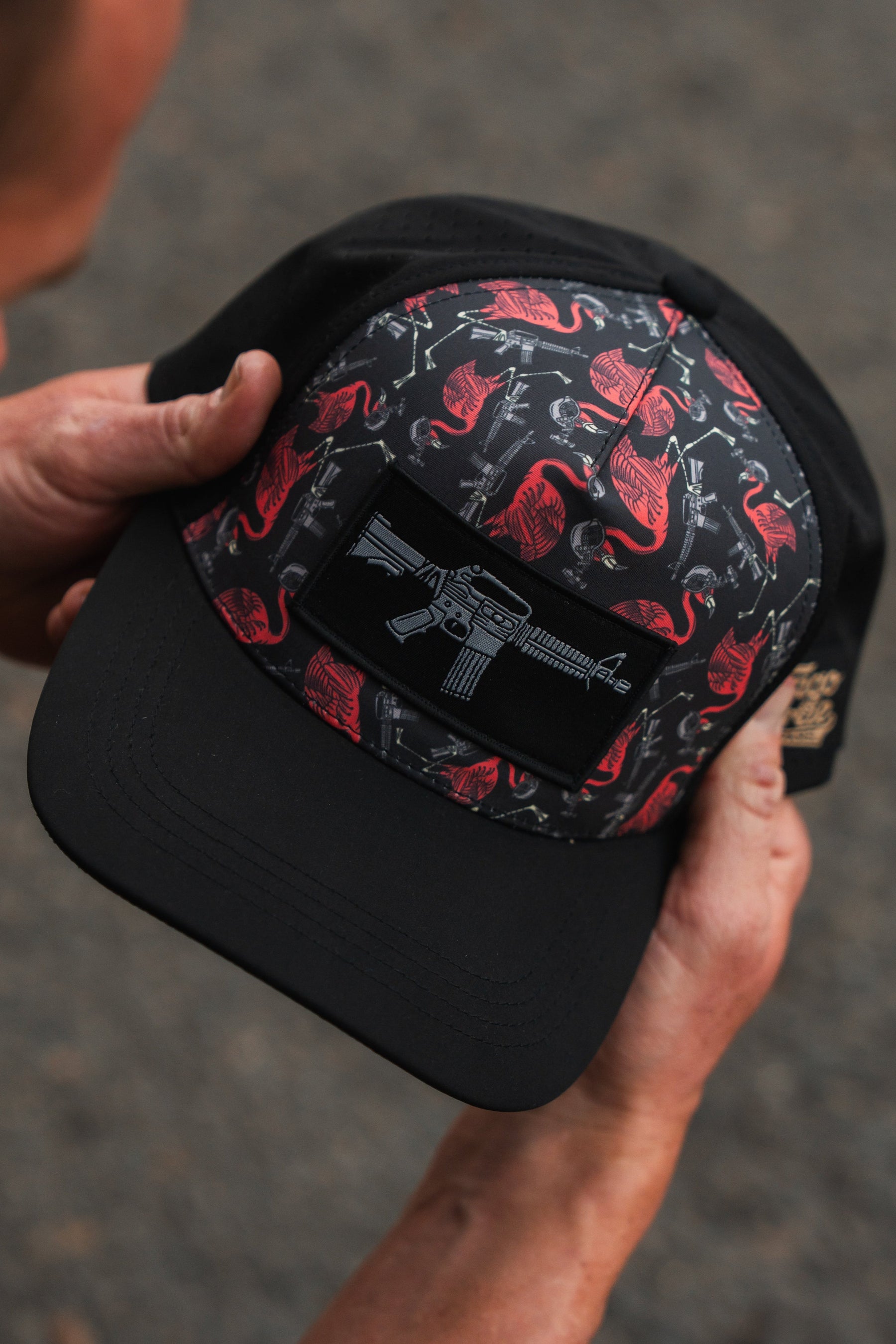Hands holding the Tango Charlie Apparel Tactimingo Hydro Snapback Hat, showcasing a sweat-resistant black cap with a red flamingo and cactus pattern, featuring a rifle patch on the front against a softly blurred outdoor scene.