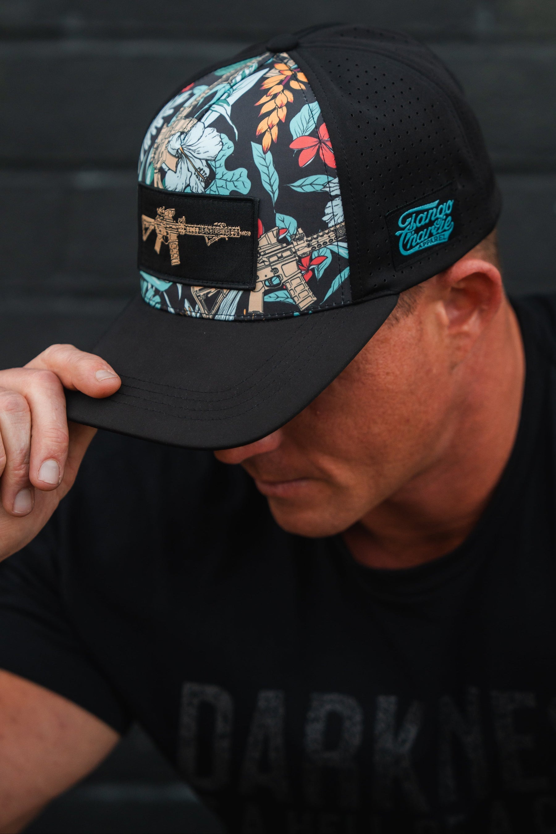 Someone wears a Tactical Tropics Hydro-Snapback Hat by Tango Charlie Apparel, showcasing colorful floral patterns and an embroidered rifle, as they tilt the snapback closure downwards against a dark, textured backdrop.
