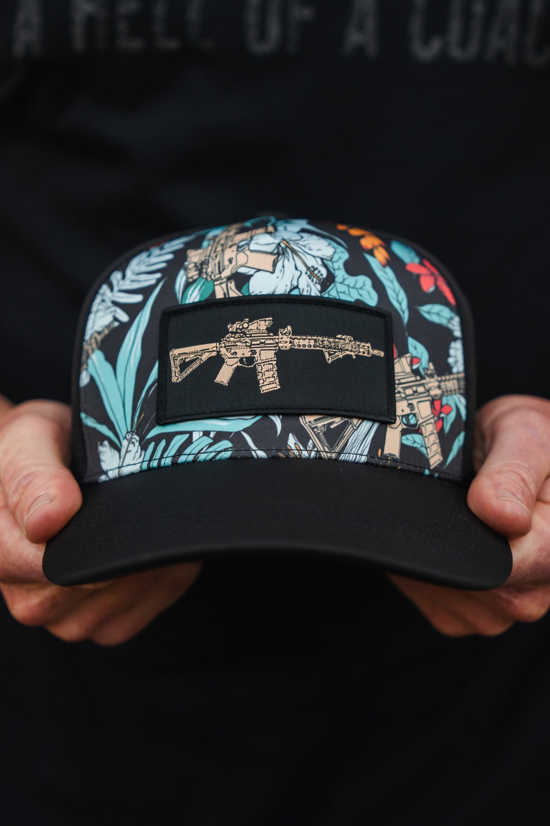 Someone is holding a Tactical Tropics Hydro - Snapback Hat from Tango Charlie Apparel. This hat features a dark, vibrant floral pattern with an embroidered rifle patch on the front and includes a snapback enclosure.