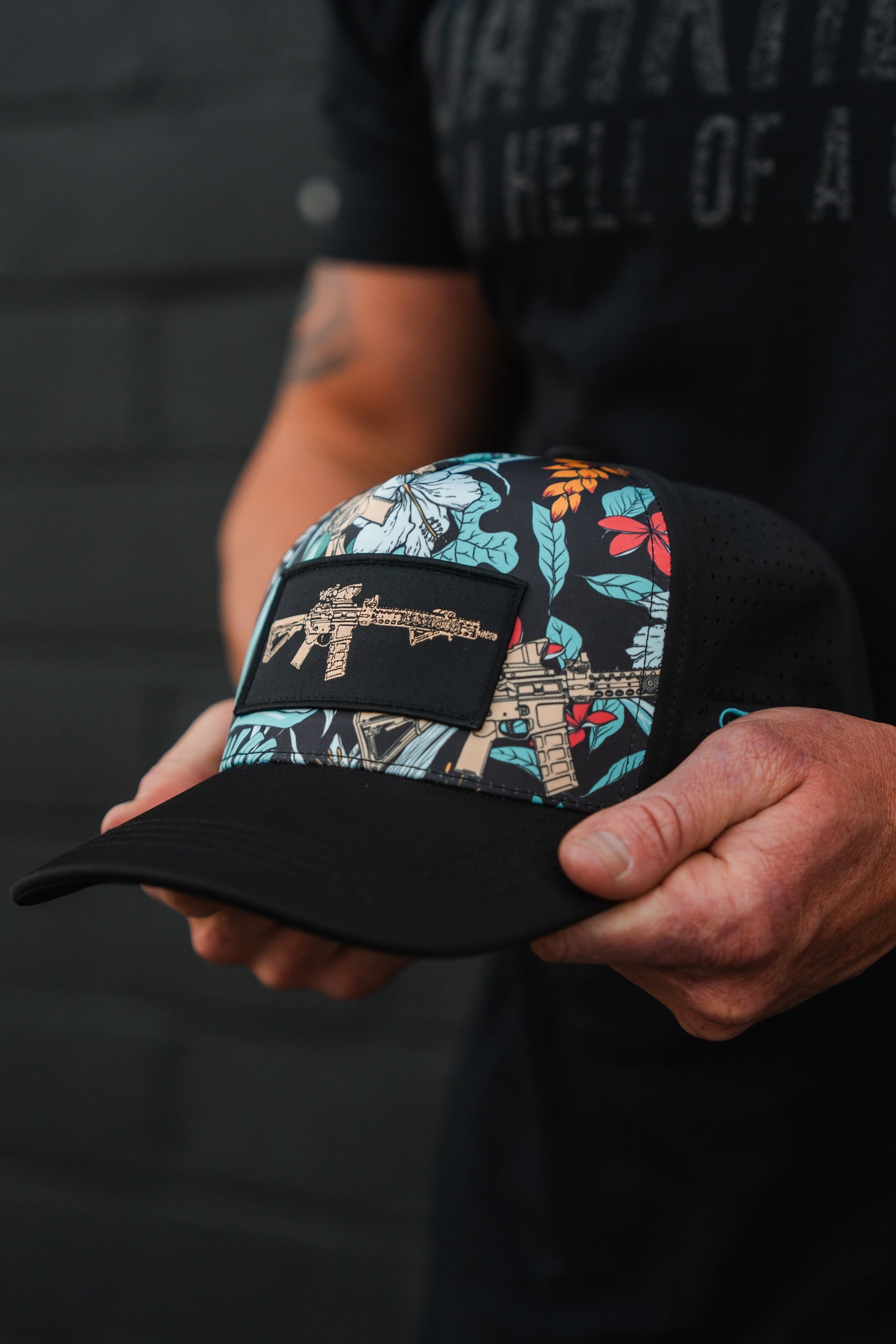 A person holds the Tango Charlie Apparels Tactical Tropics Hydro Snapback Hat, featuring a black floral design and rifle patch. Against a dark shirt and dim background, their focused hands subtly highlight the hat’s stylish hydro-resistance and snapback enclosure.