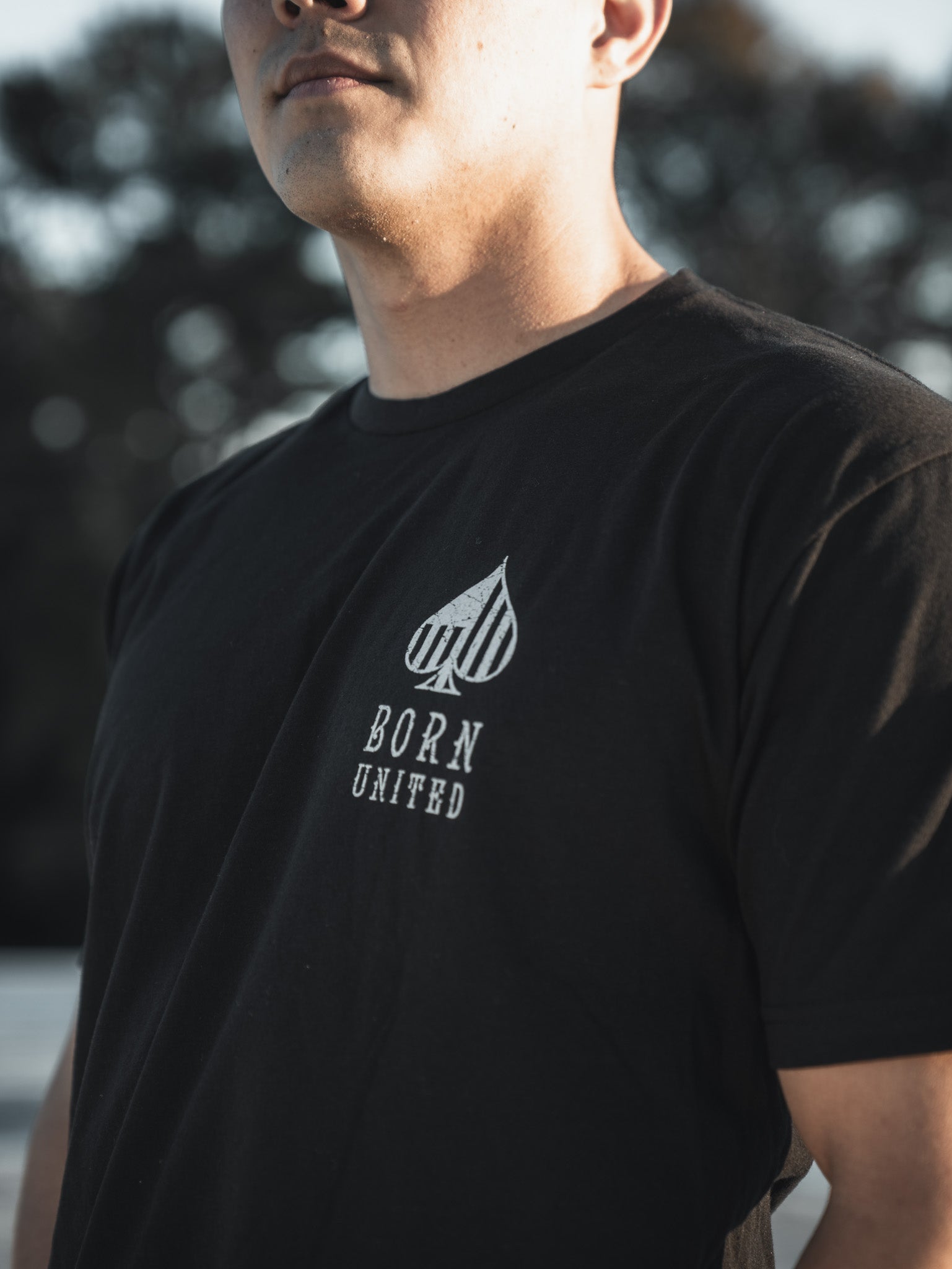 A person wearing a black Angel of Death T-shirt by Born United Private Label stands outdoors, with trees and a clear sky blurring behind them. The shirt features an upward-pointing spade and BORN UNITED. Only the torso and part of the face are visible, evoking the presence of an AC-130 aircraft above.