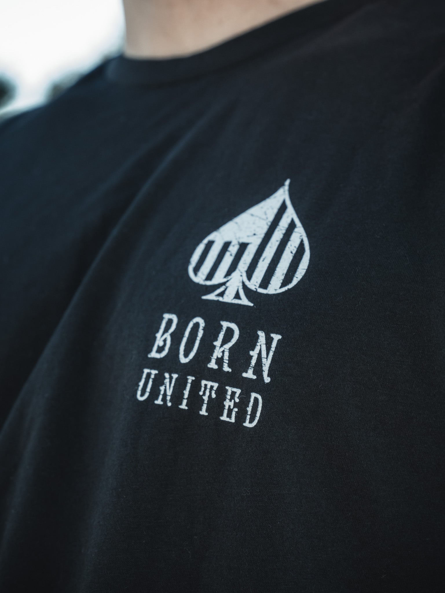 A person wears a Born United Private Label Angel of Death black T-shirt, featuring an abstract spade-like design reminiscent of an AC-130. Vertical stripes are above the distressed font words BORN UNITED.
