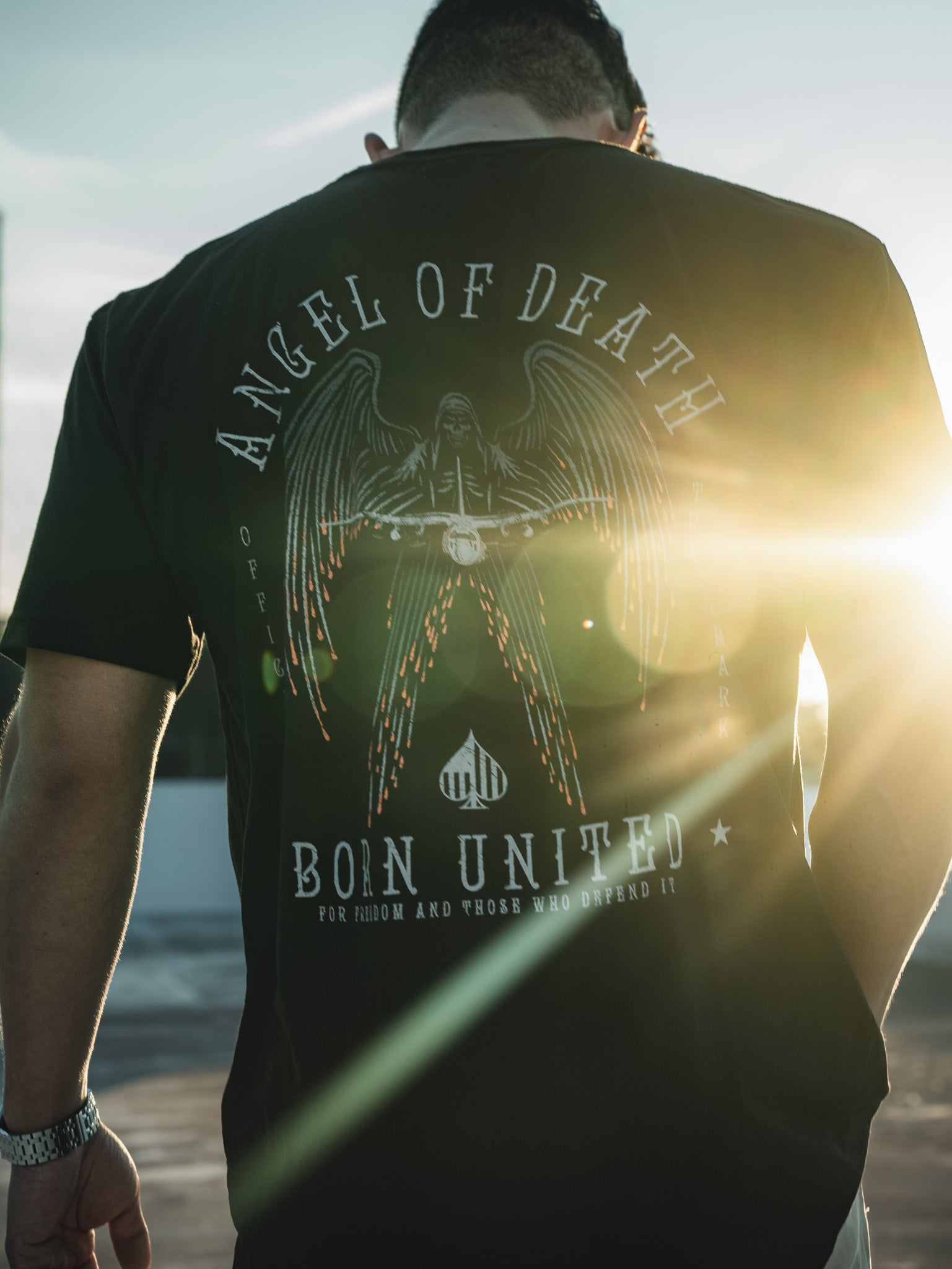 A person wearing Born United Private Labels Angel of Death black T-shirt, featuring an AC-130 military aircraft graphic under a fiery sky, stands with their back to the camera. The sunset lens flare on the right illuminates the design.