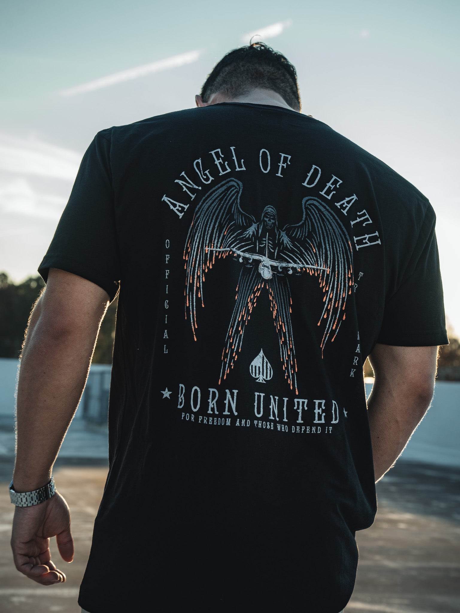 A man faces away from the camera, wearing a black Angel of Death T-shirt by Born United Private Label. The graphic shows an angel with swords, evoking the legendary AC-130 aircraft. Hes outdoors under a wide blue sky.