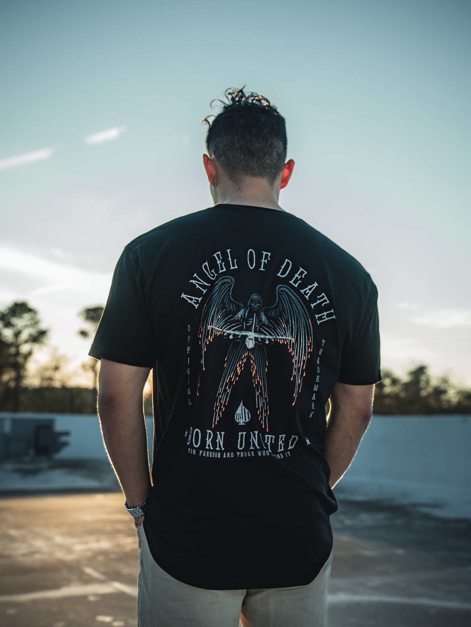 A man with curly hair stands on a rooftop, facing away and wearing a Born United Private Label black T-shirt featuring the Angel of Death design. With a clear sky and scattered clouds, it creates a serene backdrop for this striking image.