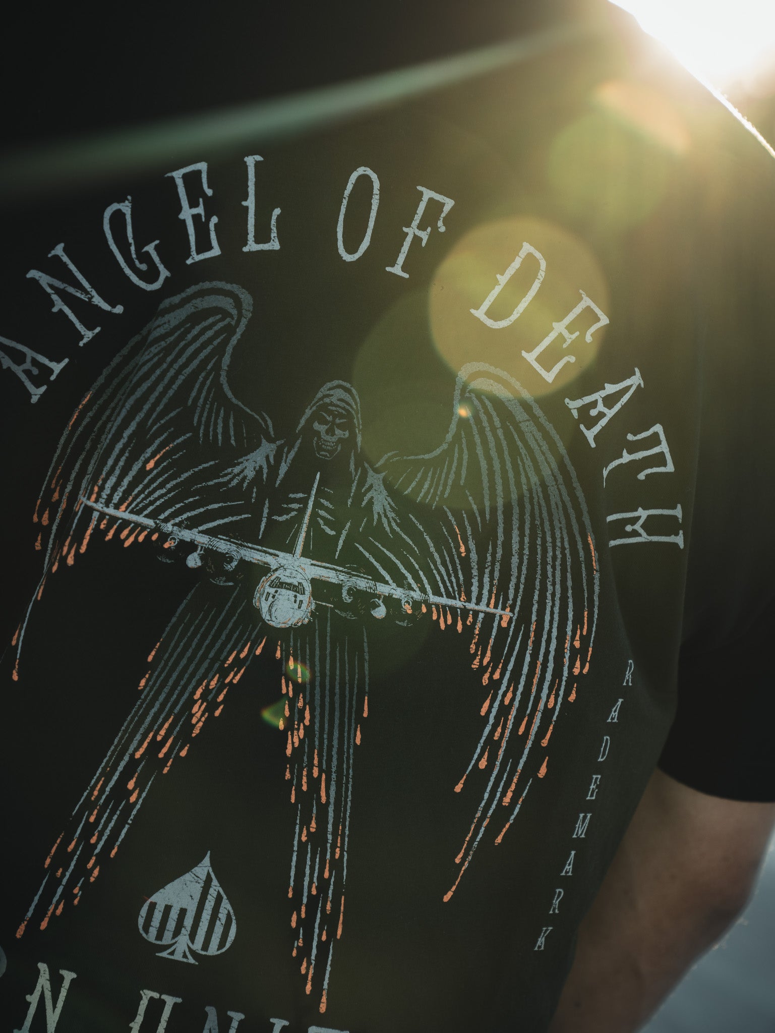 A person in a Born United Private Label black shirt displays the Angel of Death design, featuring an angel with large wings holding an AC-130 aircraft. The text ANGEL OF DEATH, TRADEMARK appears, with a backlit lens flare adding drama.