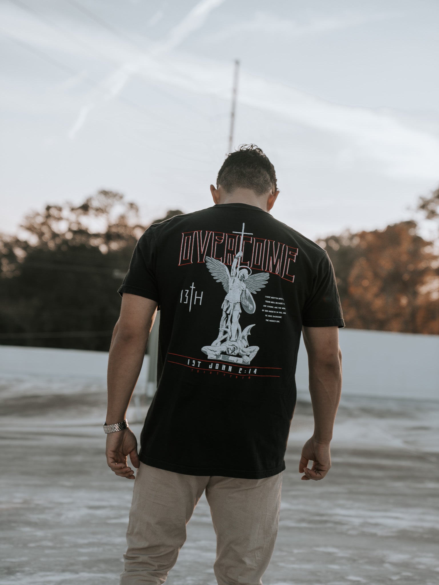 A young man stands outdoors with his back to the camera, wearing the Overcome Premium Tee by 13th. The black t-shirt features an angel graphic and Word of God, set against a serene backdrop of sunlit trees and light-colored ground.