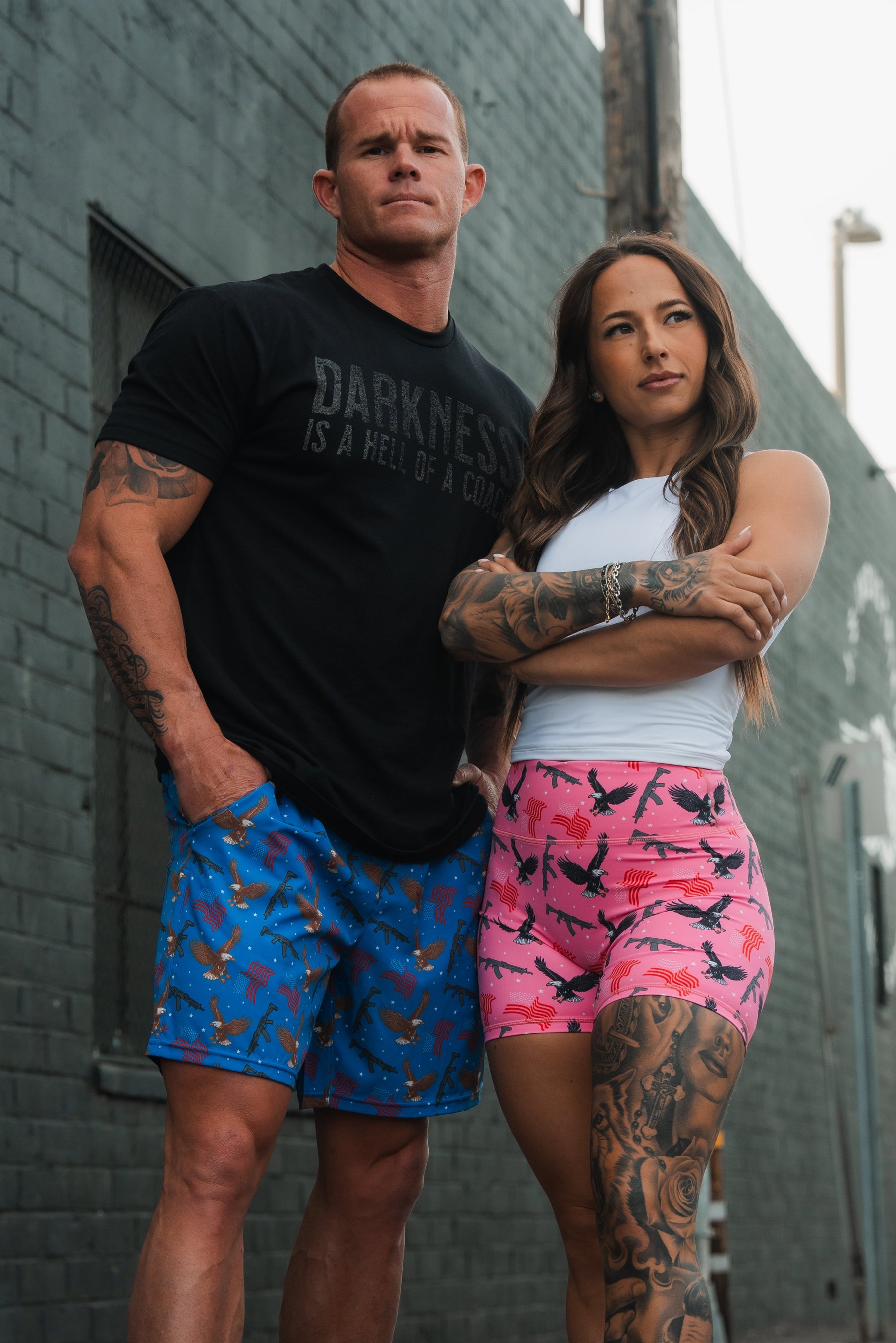 Outside, a man in a black shirt and blue shorts stands confidently beside a woman wearing a white top and Pink Victory Womens High-Waisted Seamless Front Compression Training Shorts by Born United Private Label, made of breathable fabric. Both display tattoos, exuding strength against a dark wall.