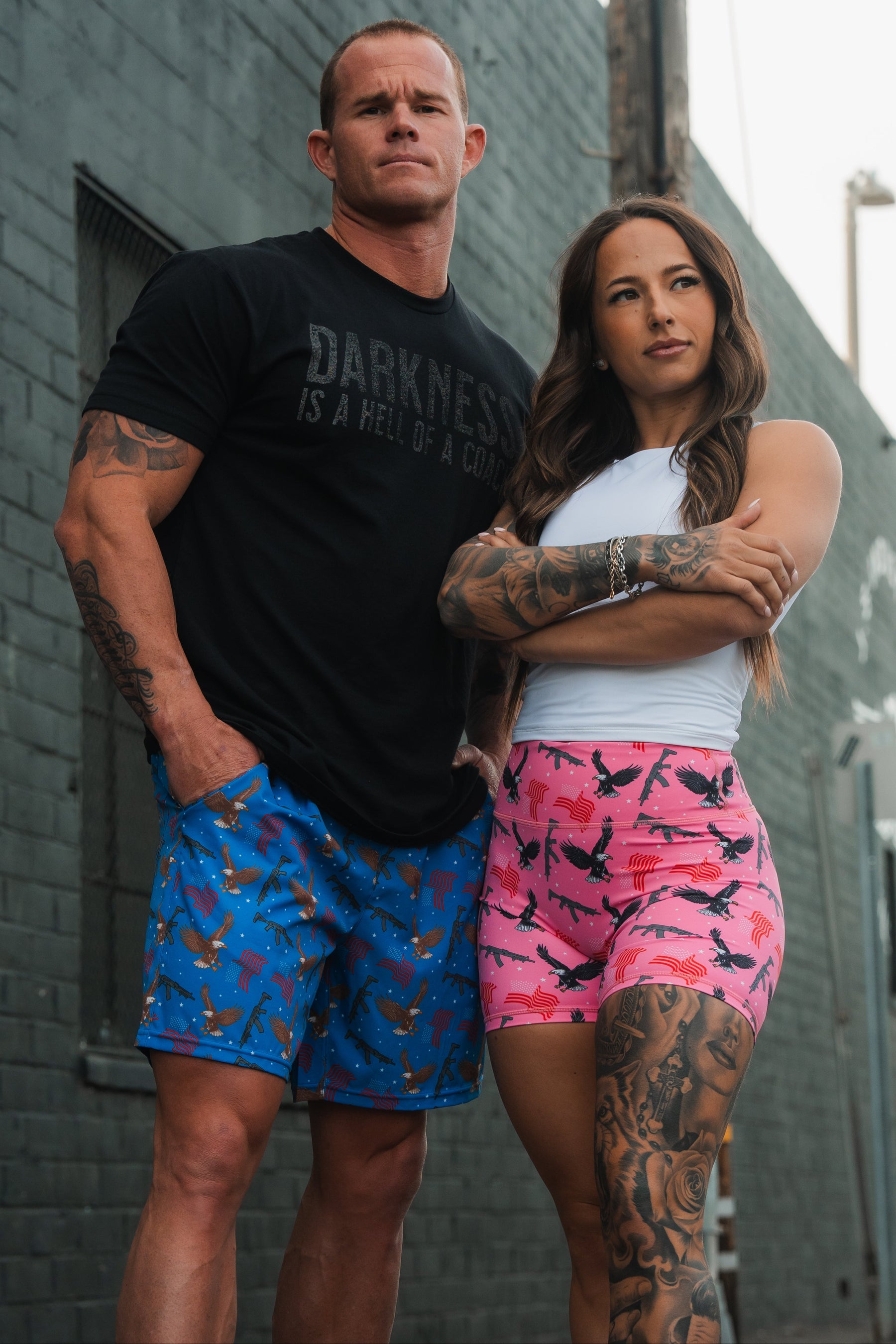 A man stands confidently in blue Mens Performance Hybrid Shorts by Born United, ideal for gym-to-swim. The woman sports a white top and pink patterned shorts. Both have tattoos, exuding presence against the urban backdrop.