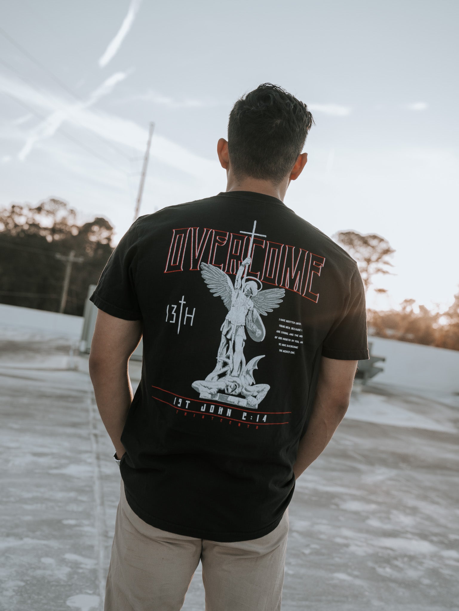 On a rooftop against a clear sky with contrails, a young man faces away wearing the Overcome Premium Tee by 13th. The black T-shirt features a large angel graphic and the words Overcome and 1st John 4:17, reflecting the word of God, with trees in the background.