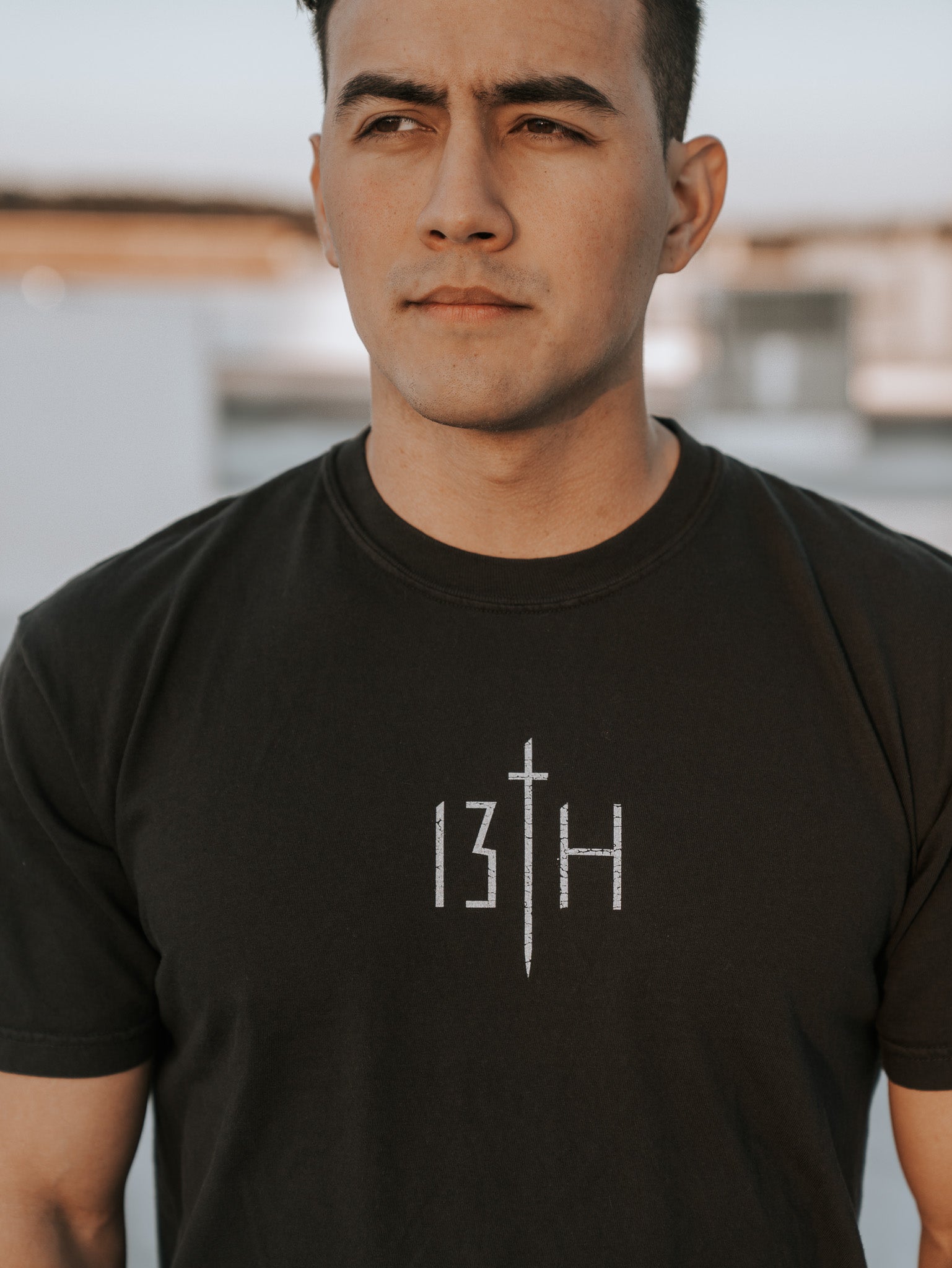A young man stands outdoors in the Overcome Premium Tee by 13th. With short dark hair and a neutral gaze, he looks into the distance, embodying resilience and the spirit to overcome. The background remains softly blurred.