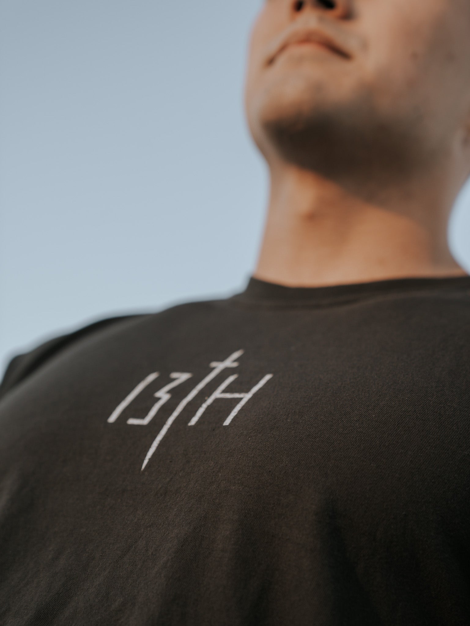 Close-up of a young man wearing the Overcome Premium Tee by 13th, a dark t-shirt featuring the brand name in white. His face is partially visible as he looks up at the clear blue sky, perhaps seeking inspiration to overcome challenges.