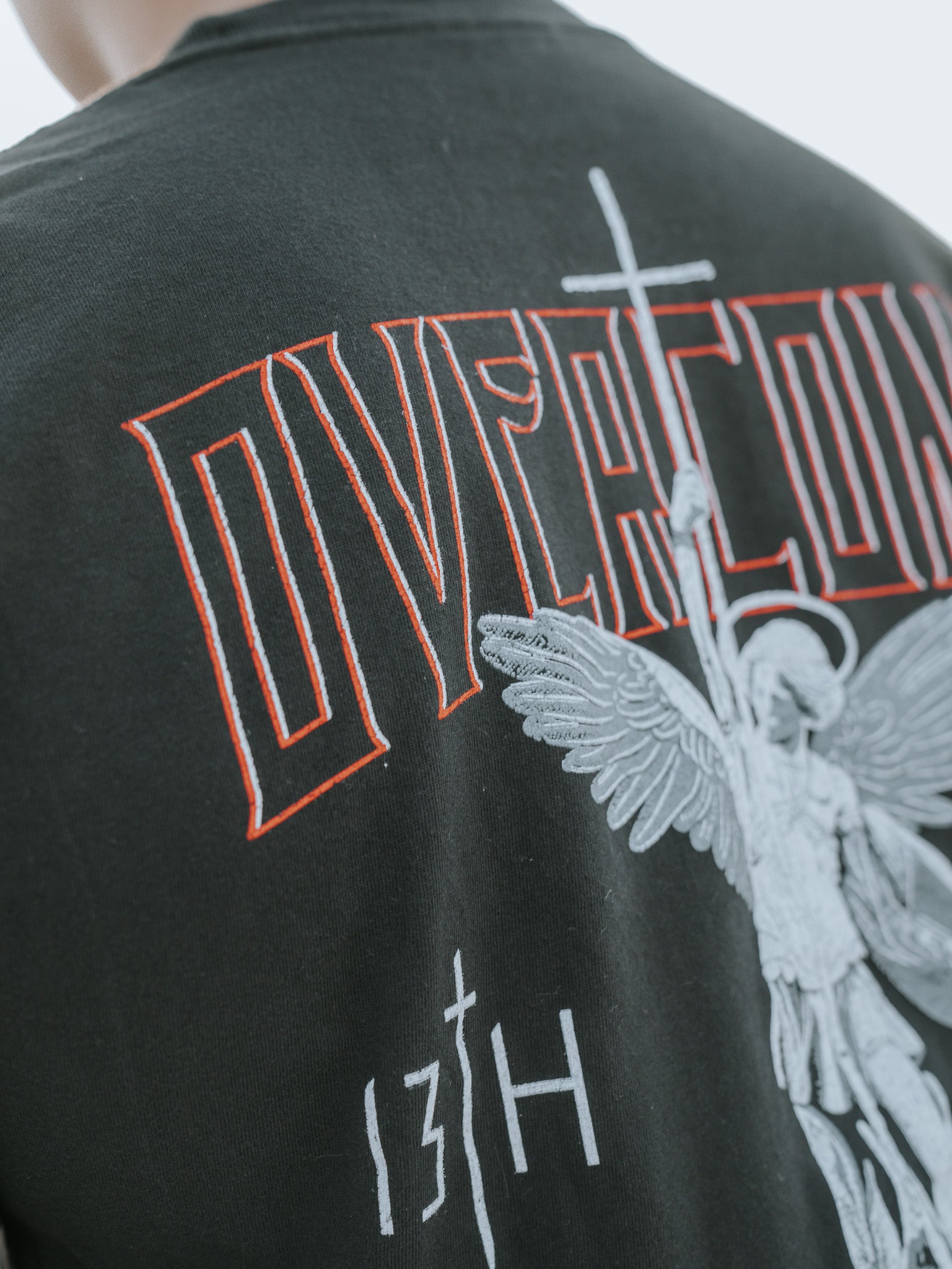 Close-up of the Overcome Premium Tee by 13th, a black shirt for young men. It features an angel graphic holding a spear and cross, with OVERCOME in red above and 13TH below, symbolizing strength through Gods word.