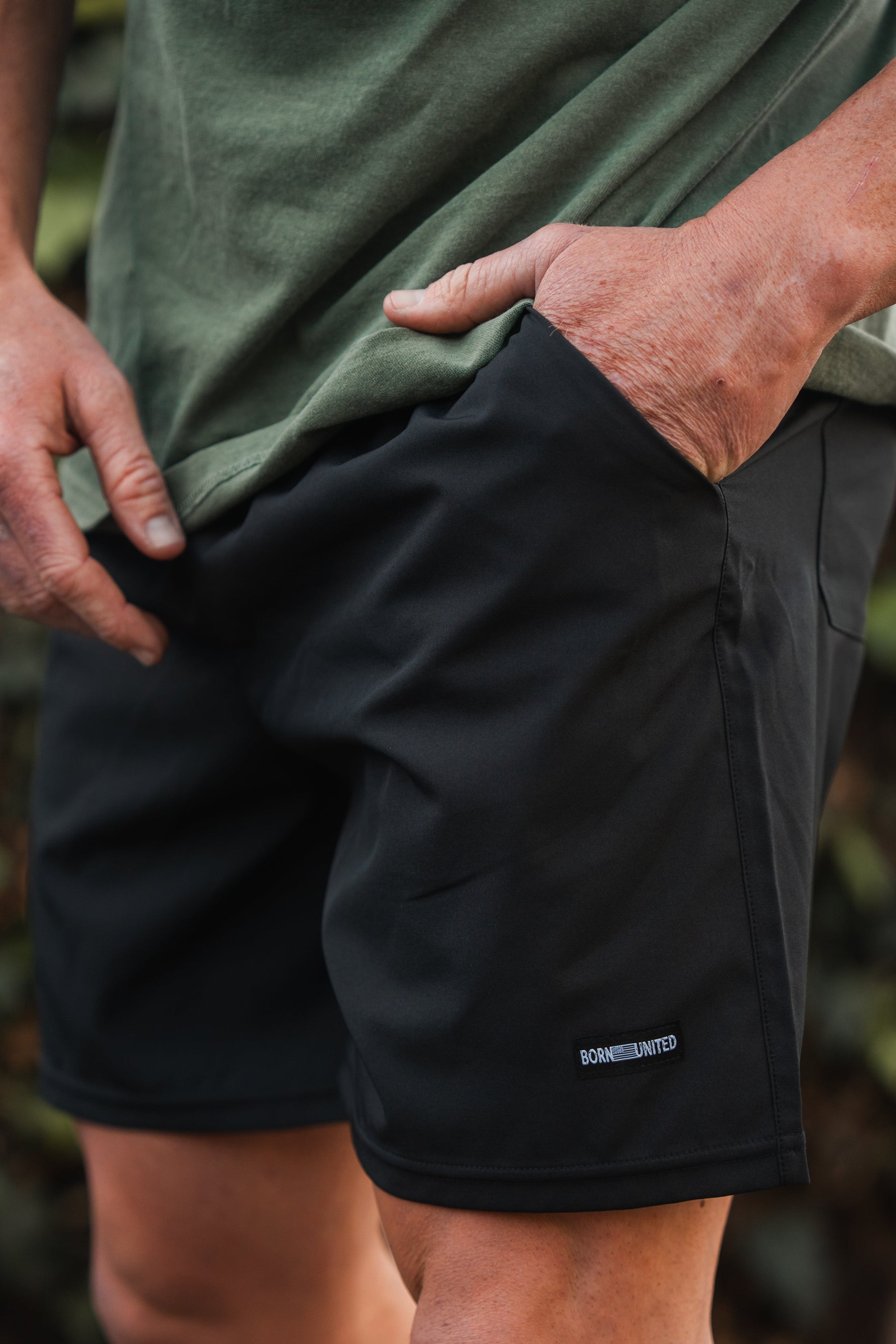 A person in a green shirt and black Mens Standard Performance Shorts | 5.5- Stealth by Born United Private Label stands outdoors, with one hand in a pocket showcasing the breathable, stretchable material and a logo near the hem.