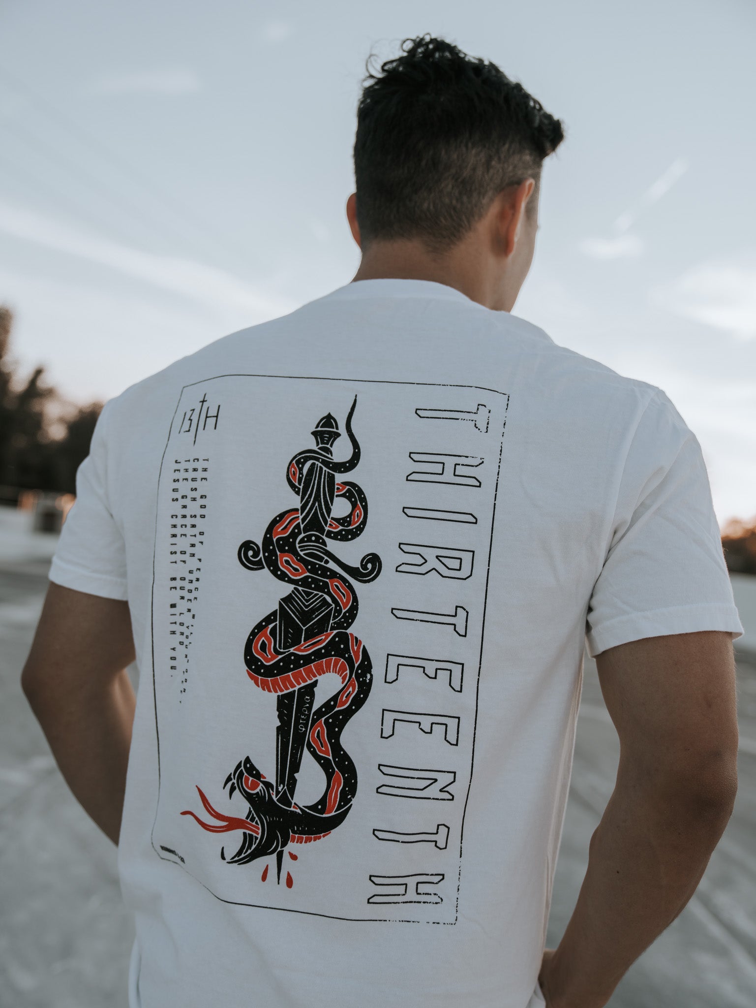 A person outdoors dons a white Lucifers Demise Premium Tee by 13th, featuring a graphic of a sword with an entwined snake symbolizing Crush Satan, and the word THIRTEENTH vertically along the side, set against a serene clear sky backdrop.