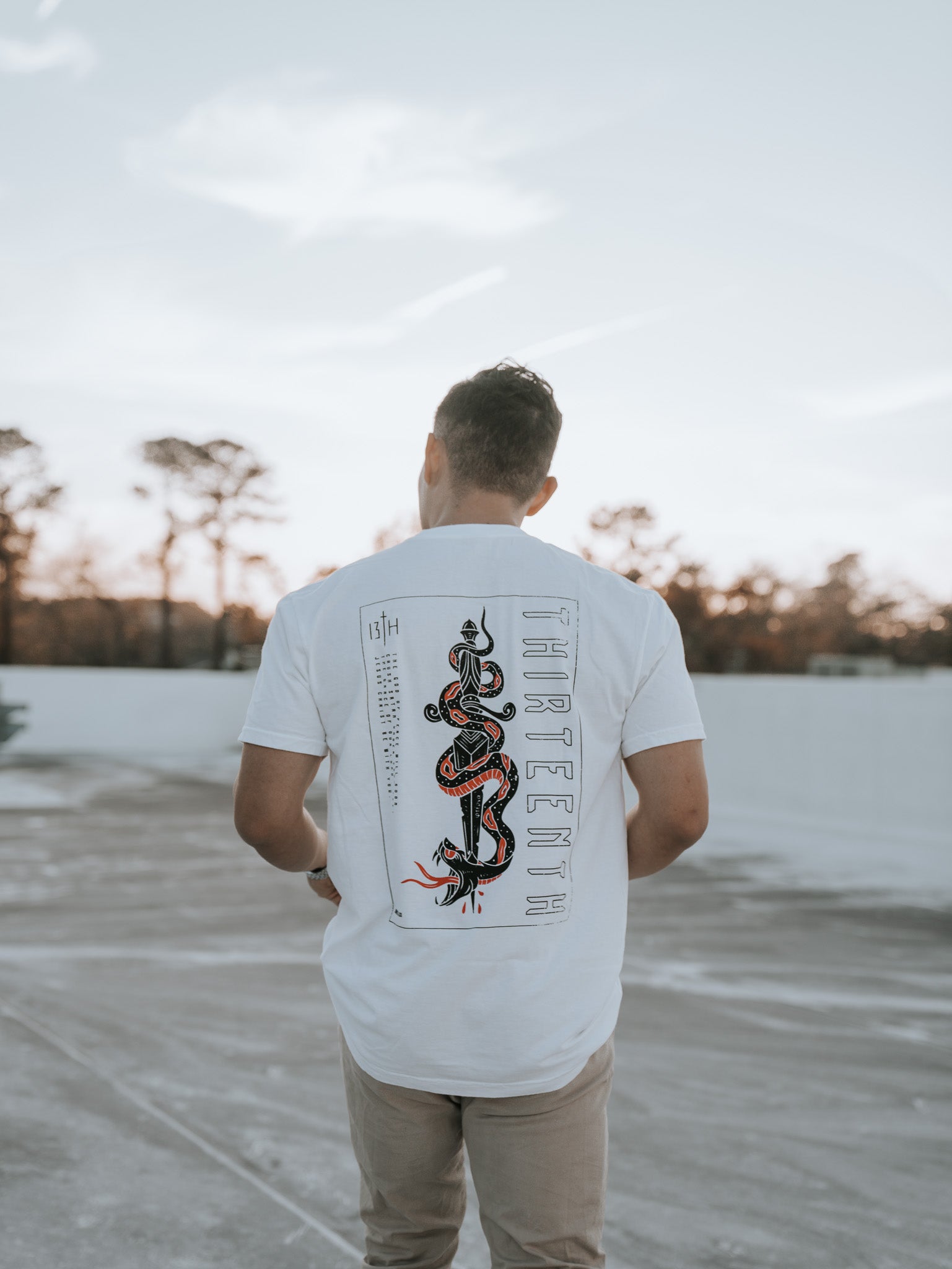 A man with short hair stands on a concrete surface outdoors, facing away. Hes wearing a 13th brand Lucifers Demise Premium Tee in white featuring a snake graphic and the words Crush Satan on the back. The sky is clear with trees in the serene background.
