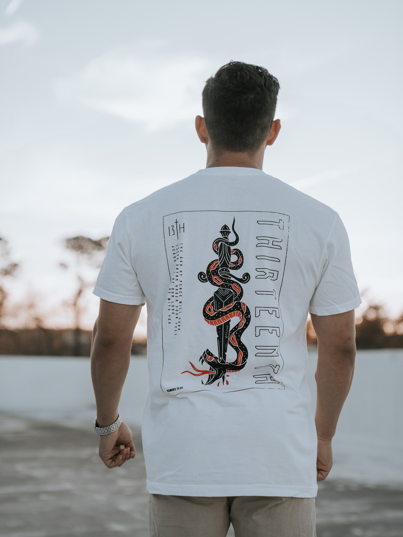 A person with short hair stands outside, wearing the 13th brand Lucifers Demise Premium Tee in white, featuring a red and black snake and THREEFOUR on the back. The clear sky and gently swaying distant trees create a serene scene.