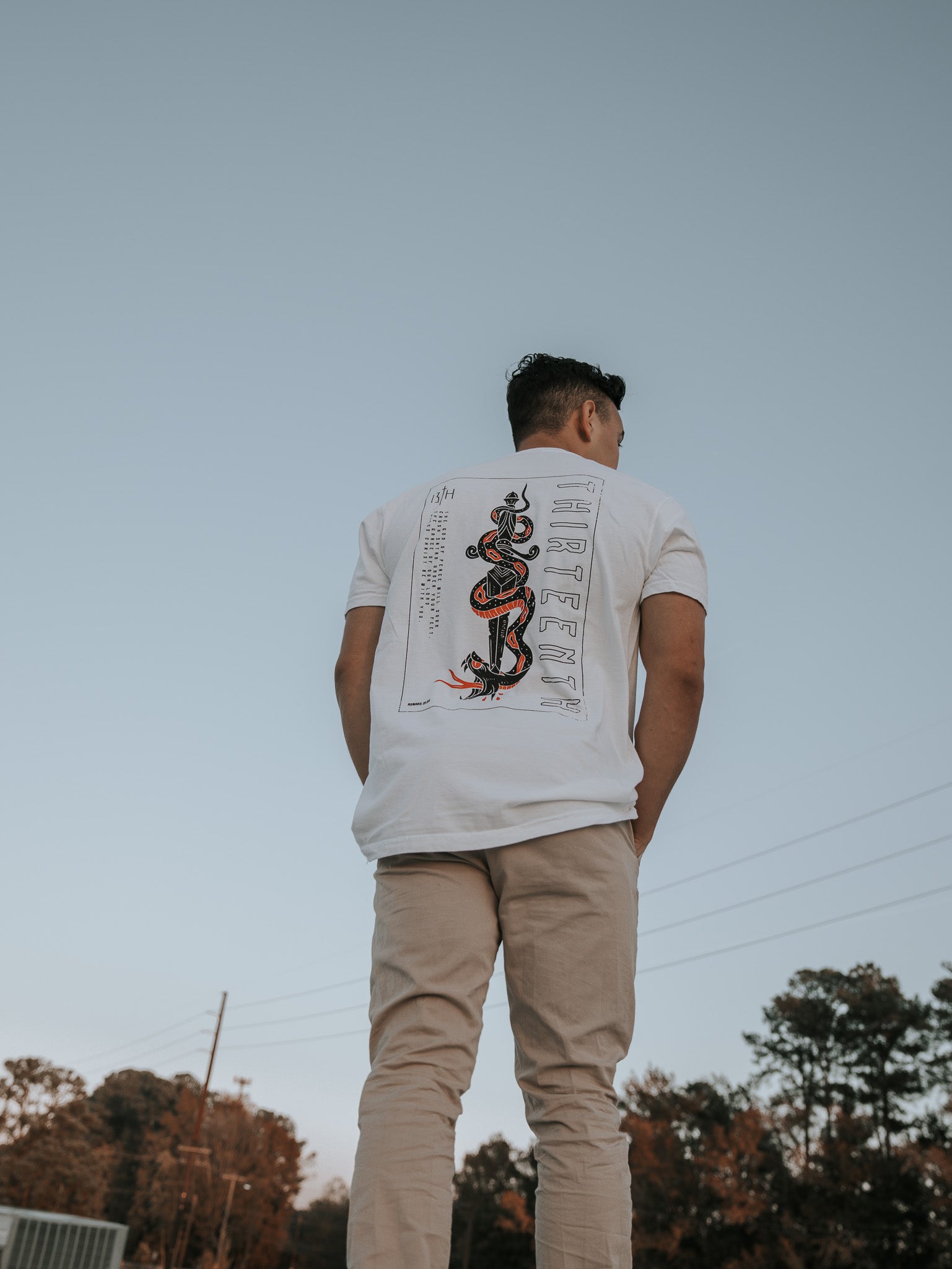 A man wearing the 13ths Lucifers Demise Premium Tee- White with a snake and Crush Satan graphic stands outdoors in beige pants, set against a clear sky with trees and power lines providing a serene background.
