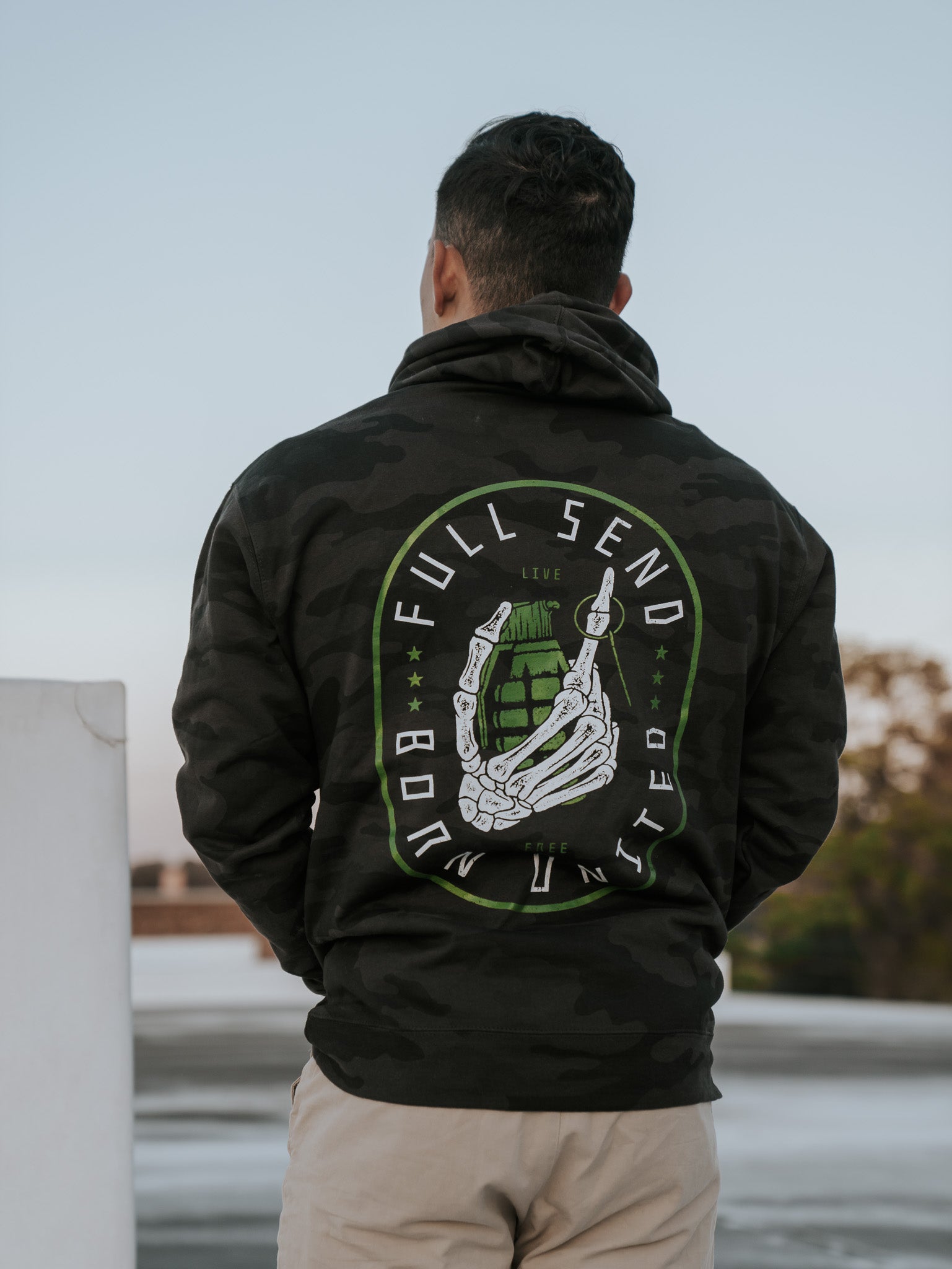 A person stands with their back to the camera, wearing the Born United Private Label Full Send Camo Hoodie (Warehouse) with a striking skeleton hand and grenade design. Theyre outdoors against a blurred backdrop of trees and buildings.