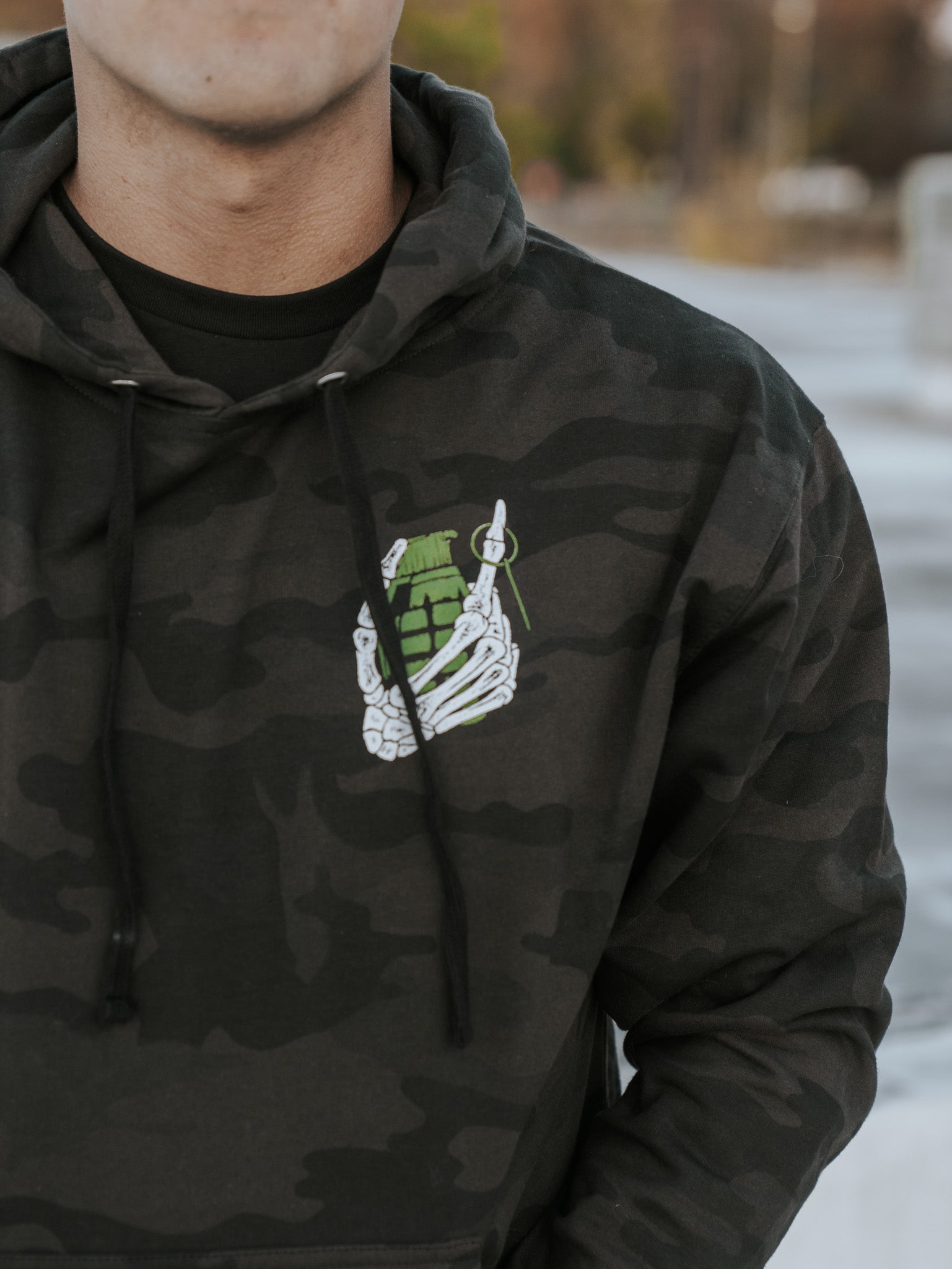 Wearing the Born United Private Labels Full Send Camo Hoodie (Warehouse), featuring a skeletal hand holding a green grenade, the person exudes the Full Send vibe with their face hidden against a blurred outdoor background.