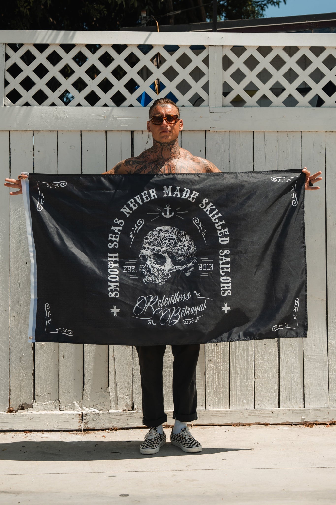 Outdoors, a tattooed man holds the Skilled Sailor Flag by Relentless Betrayal, featuring a pirate skull and the text Smooth seas never made skilled sailors and Relentless Barangay. Wearing sunglasses, a pendant, checkered shoes, and black trousers, he captures the spirit of resilience.