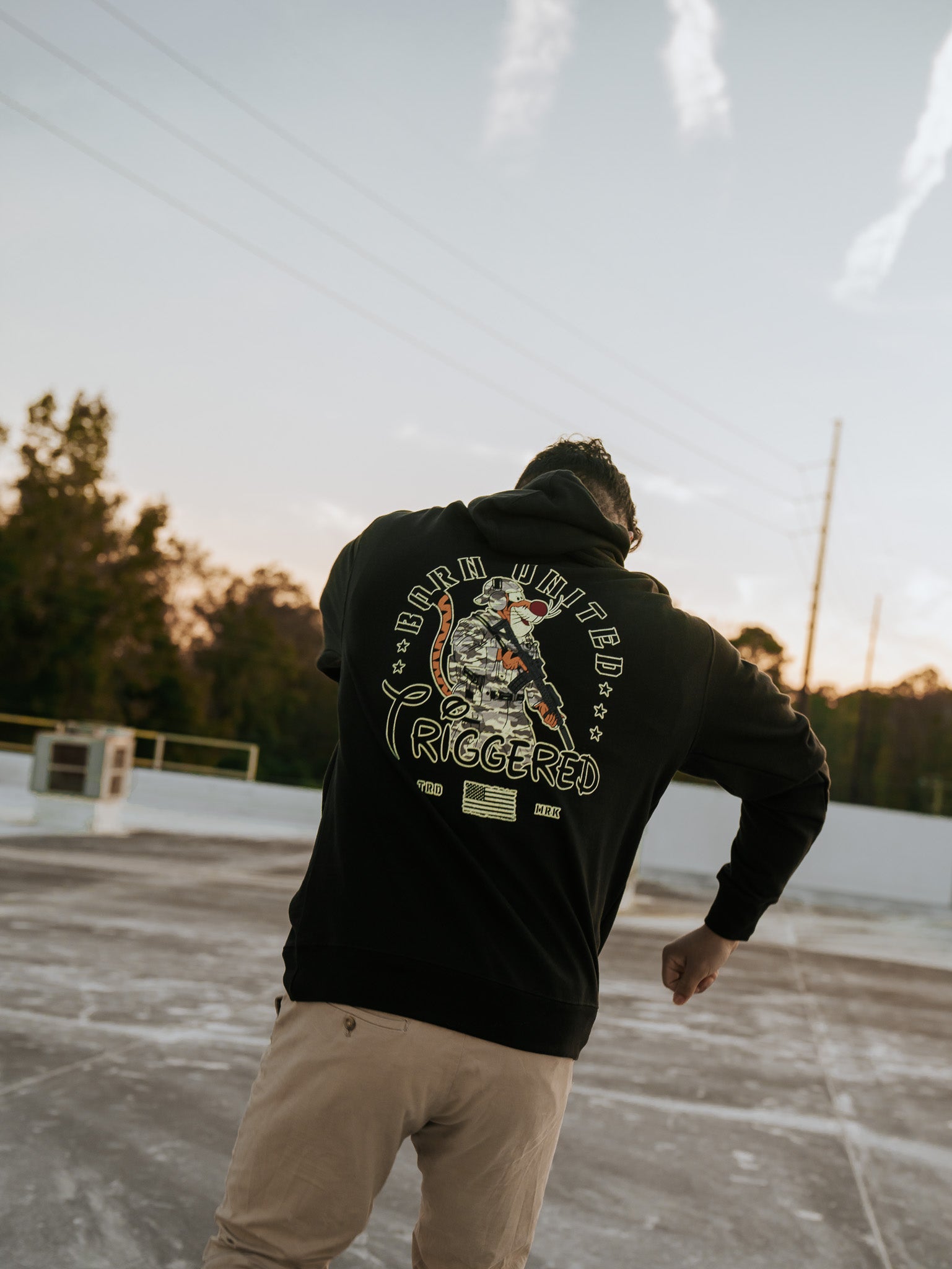 A person wearing the Triggered Hoodie by Born United Private Label, featuring graphic designs of a childhood character and symbols, stands on a concrete surface with trees and a sunset sky in the background as they look away from the camera.
