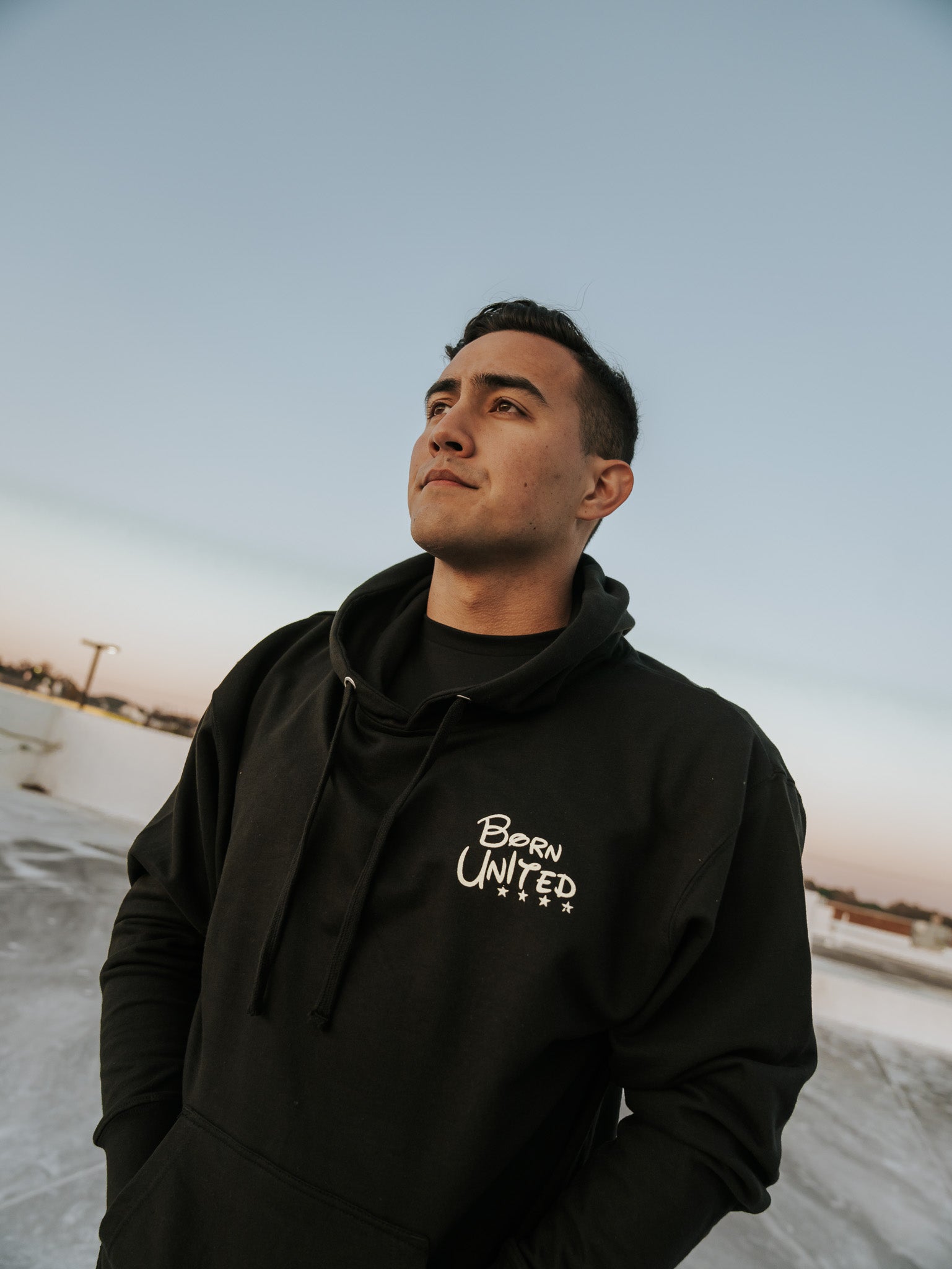 A person gazes into the distance, embodying a childhood character while wearing the Triggered Hoodie by Born United Private Label. The serene sunset sky in the backdrop transitions from blue to warm hues.