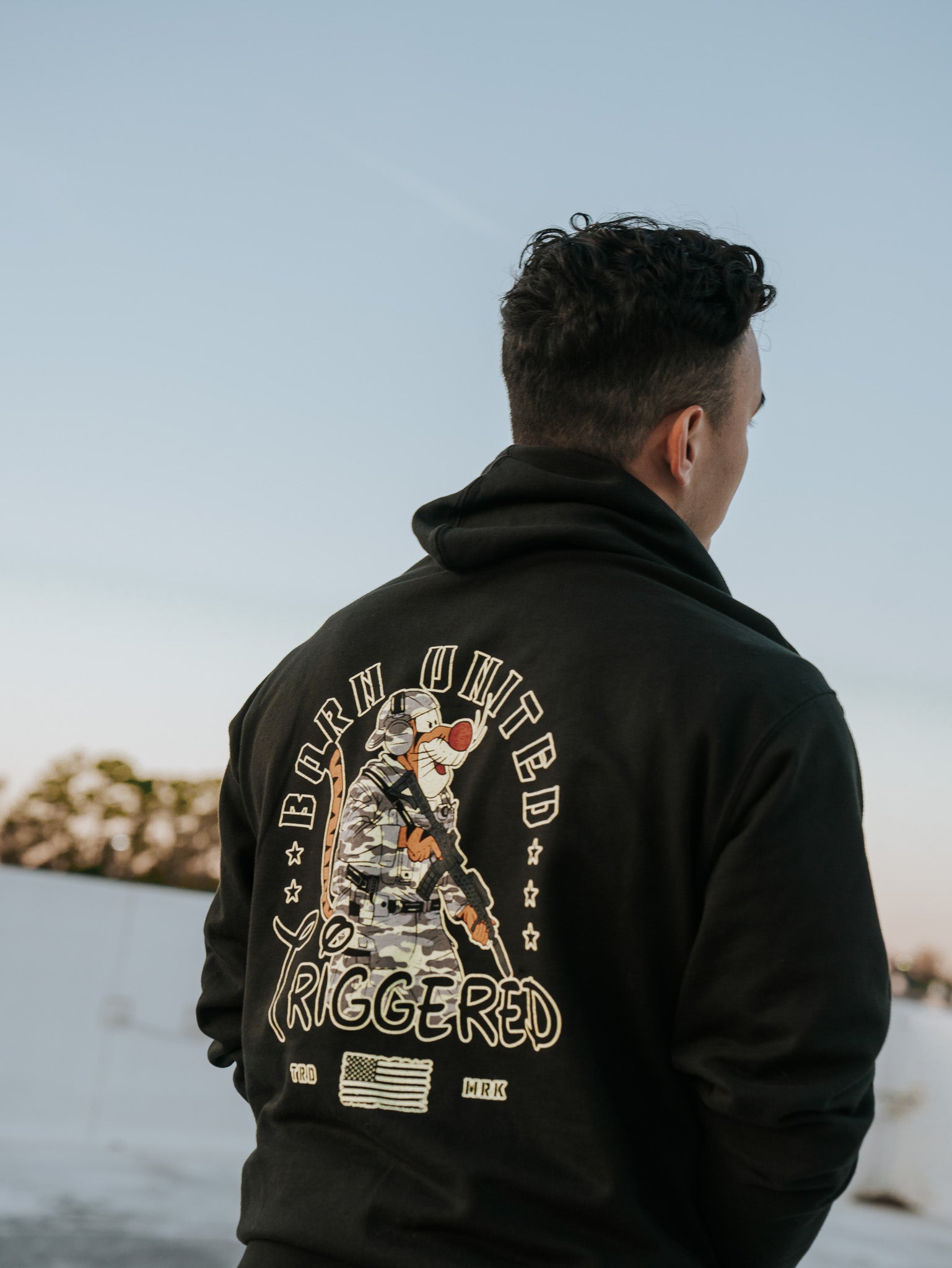 Facing away outdoors, a person with short curly hair wears the Born United Private Labels black Triggered Hoodie featuring a soldier graphic, Born United Triggered text, and a small American flag, perfectly capturing the locked and loaded spirit against the clear blue sky.
