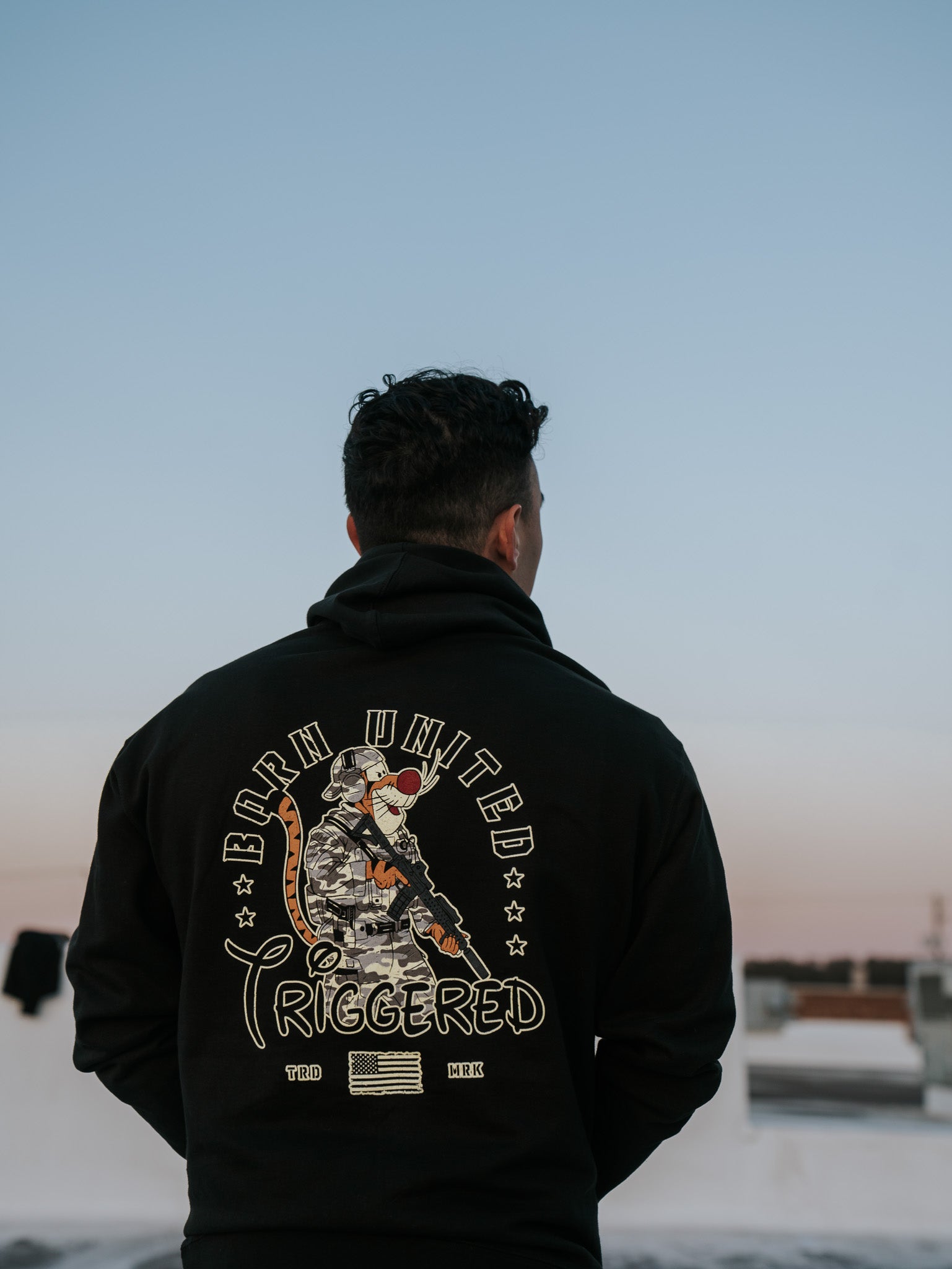 A person seen from behind wears the Triggered Hoodie by Born United Private Label, featuring a soldier graphic and the phrase Born United Triggered. The sky showcases a vibrant blue-to-orange gradient, adding an American update flair.