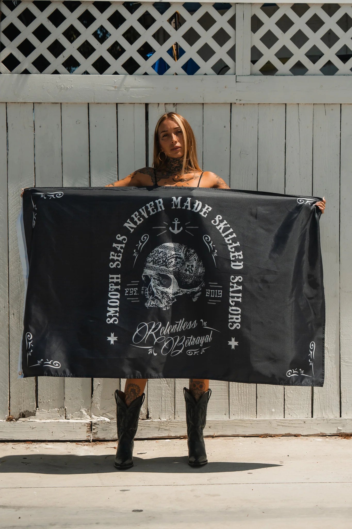 A person stands by a white wooden fence holding the Skilled Sailor Flag, a black flag featuring a skull, anchor, and the phrase Smooth Seas Never Made Resilient People, with decorative elements by Relentless Betrayal paying tribute to brave sailors.