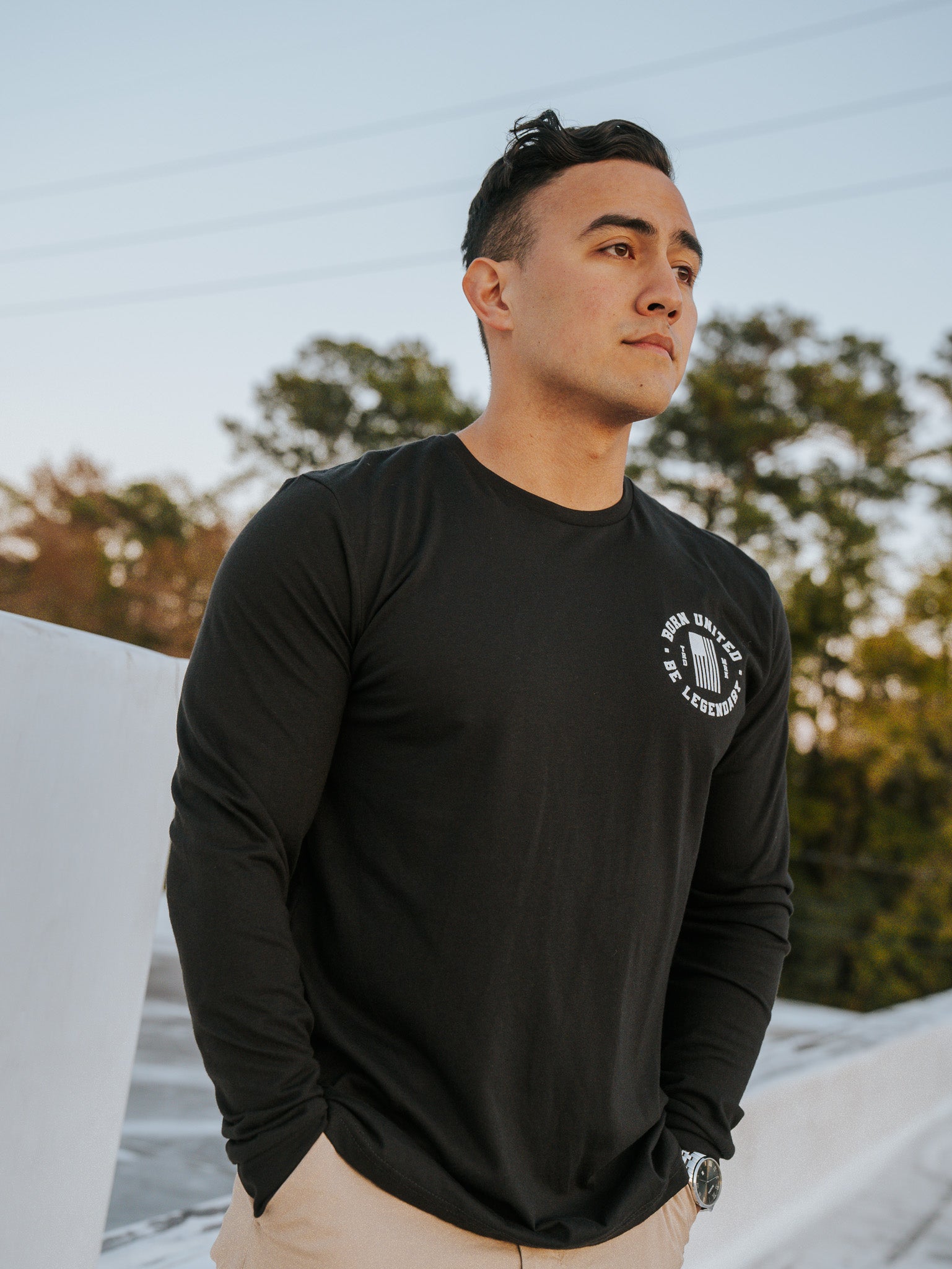 A person stands outdoors on a bridge, donning the Forever Legend Trump - Long Sleeve by Born United Private Label with beige pants, hands in pockets. The picturesque background of trees and a sunset sky enhances the serene atmosphere.