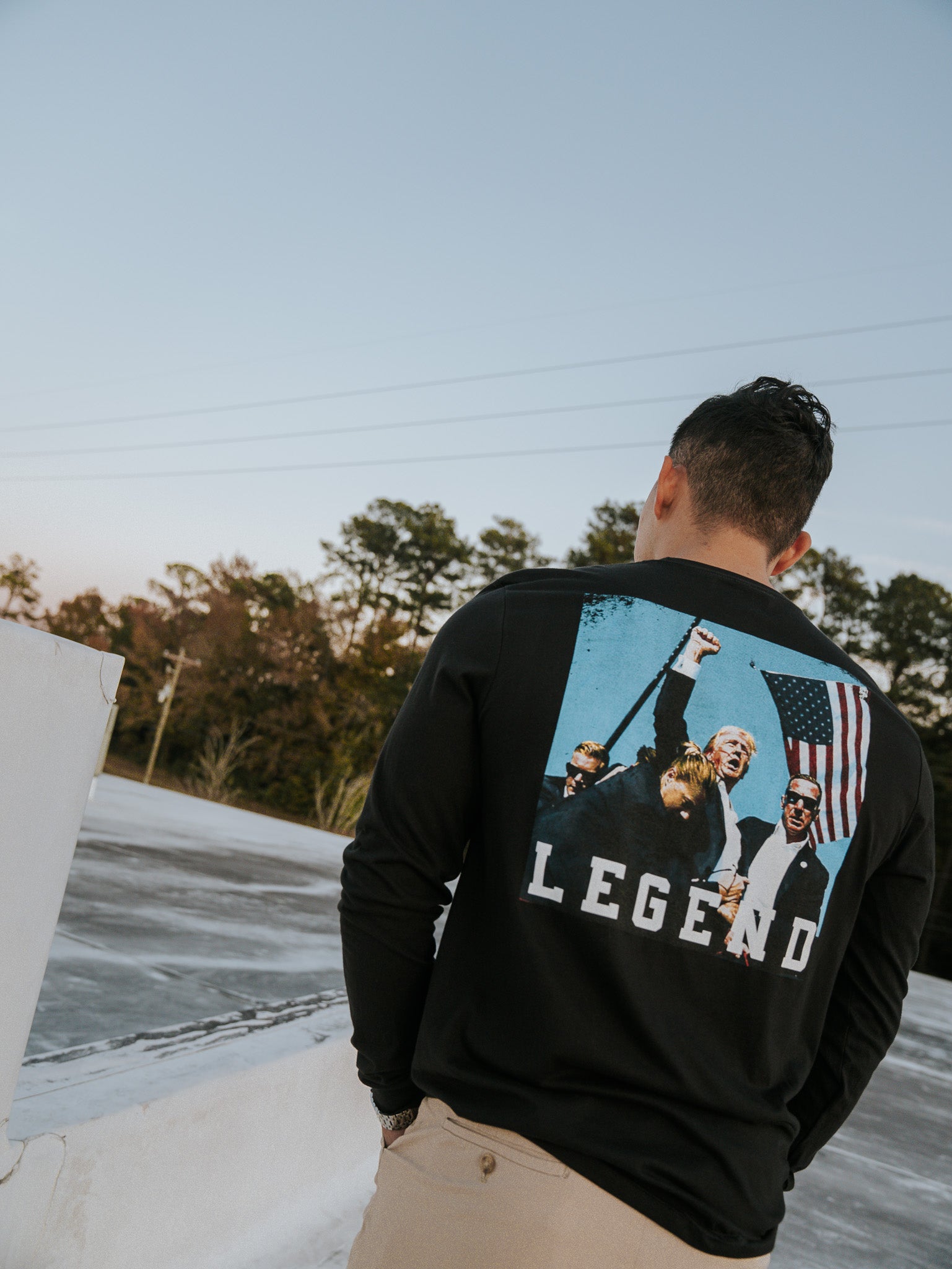 A person outdoors wears a Born United Private Label Forever Legend Trump long-sleeve shirt featuring several people, an American flag, and Forever Legend text. The vivid design is set against trees and a clear sky.