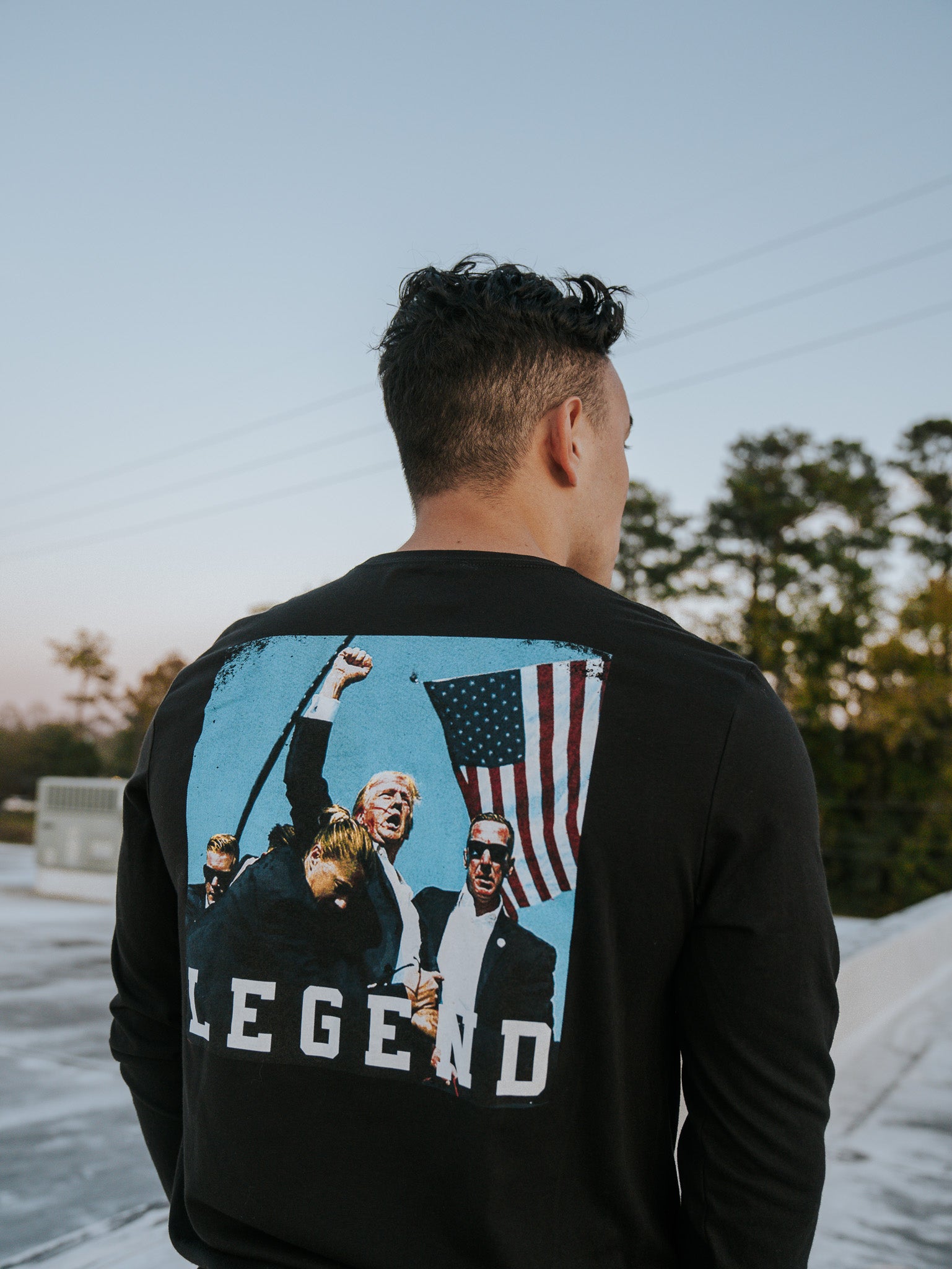 A person with short curly hair, seen from behind, wears the Born United Private Label’s Forever Legend Trump long sleeve featuring people holding an American flag and raising fists. Forever Legend is inscribed below. The background shows trees against a clear sky.