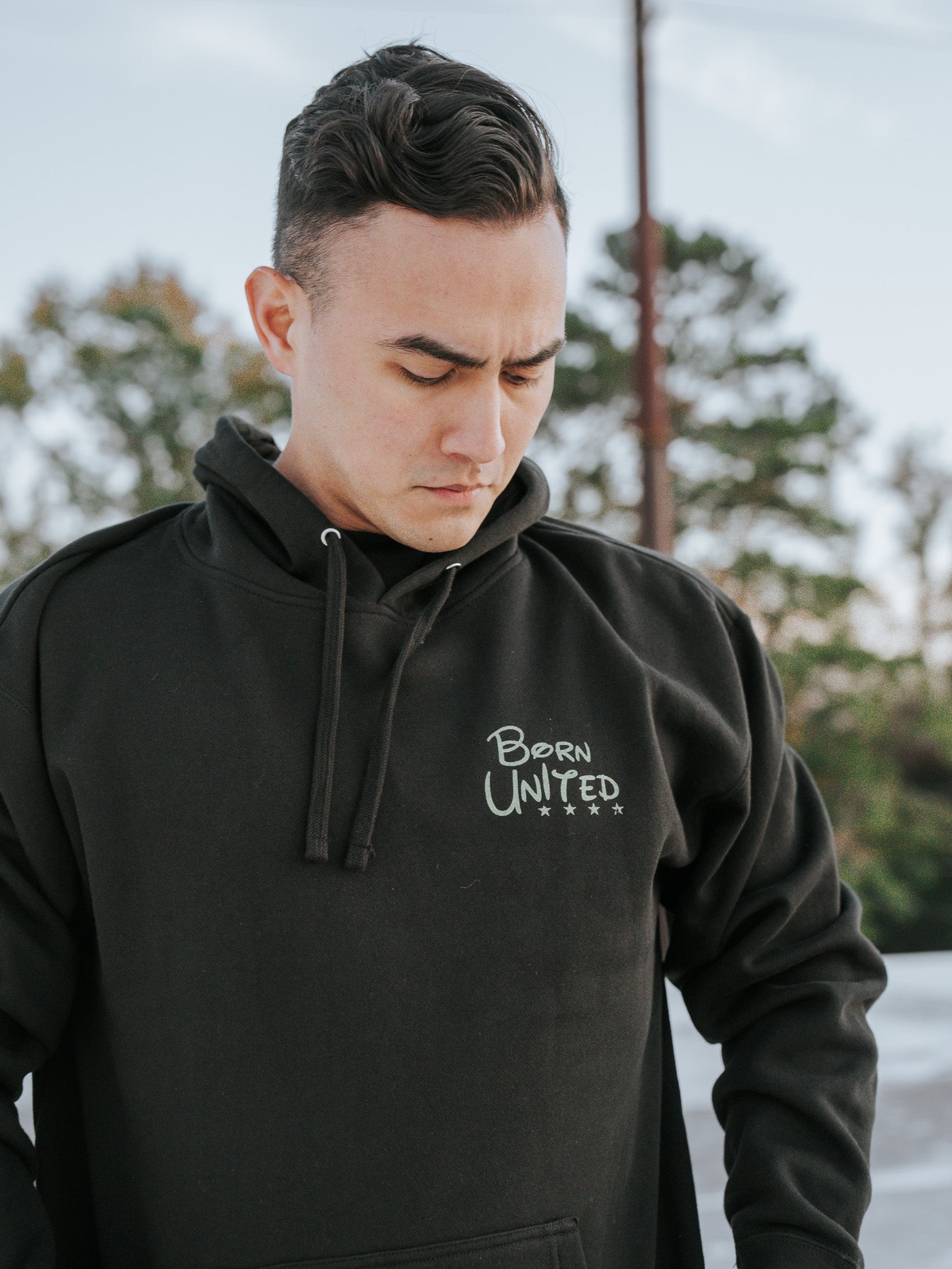 A person outdoors is wearing a black Winnie The Pew Hoodie from Born United Private Label, exuding chilled vibes amidst natures beauty. With short dark hair and a relaxed expression, they look down while surrounded by trees and a clear sky.