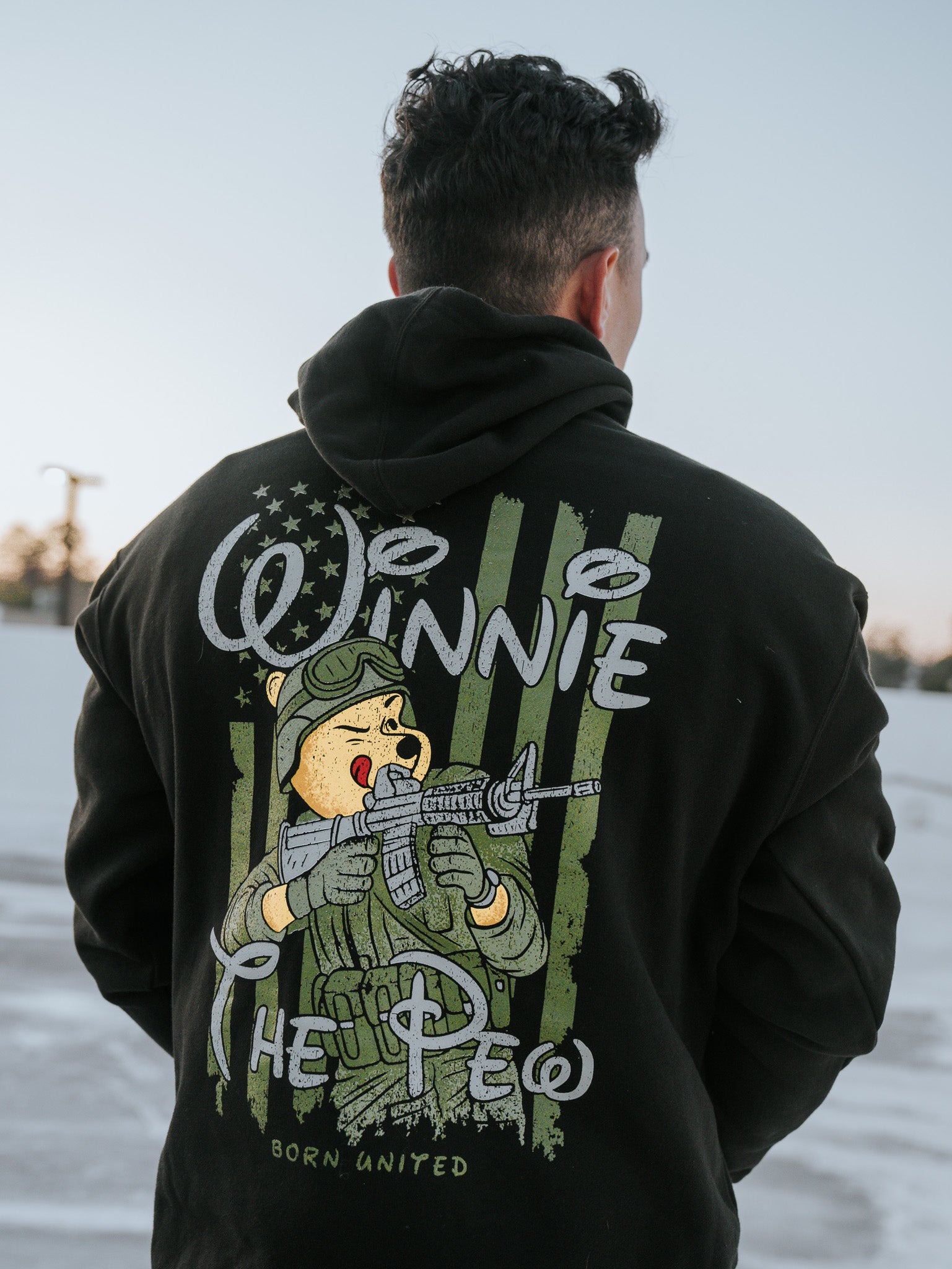 A person wears the Winnie The Pew Hoodie by Born United Private Label, showcasing a bear with a rifle and snowy scenery in the backdrop, offering whimsical style inspiration for fans of clever home decor designs.