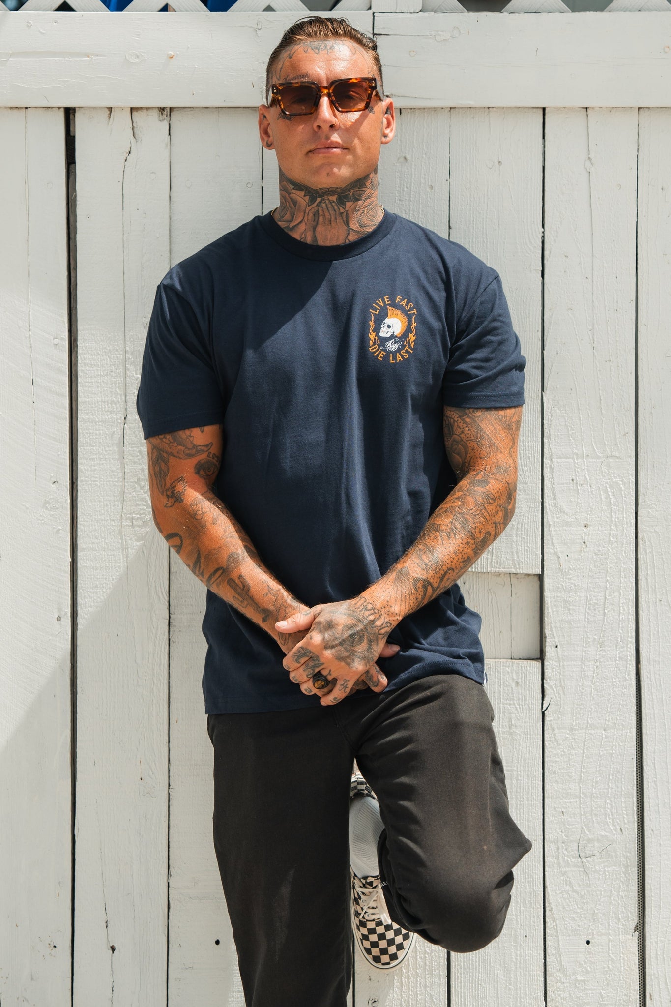 Leaning casually against a white wooden fence with one leg raised, a youthful person with tattoos and sunglasses radiates adventure, hands clasped in a relaxed pose. Theyre dressed in black pants and the Live Fast Navy T-Shirt by Relentless Betrayal.