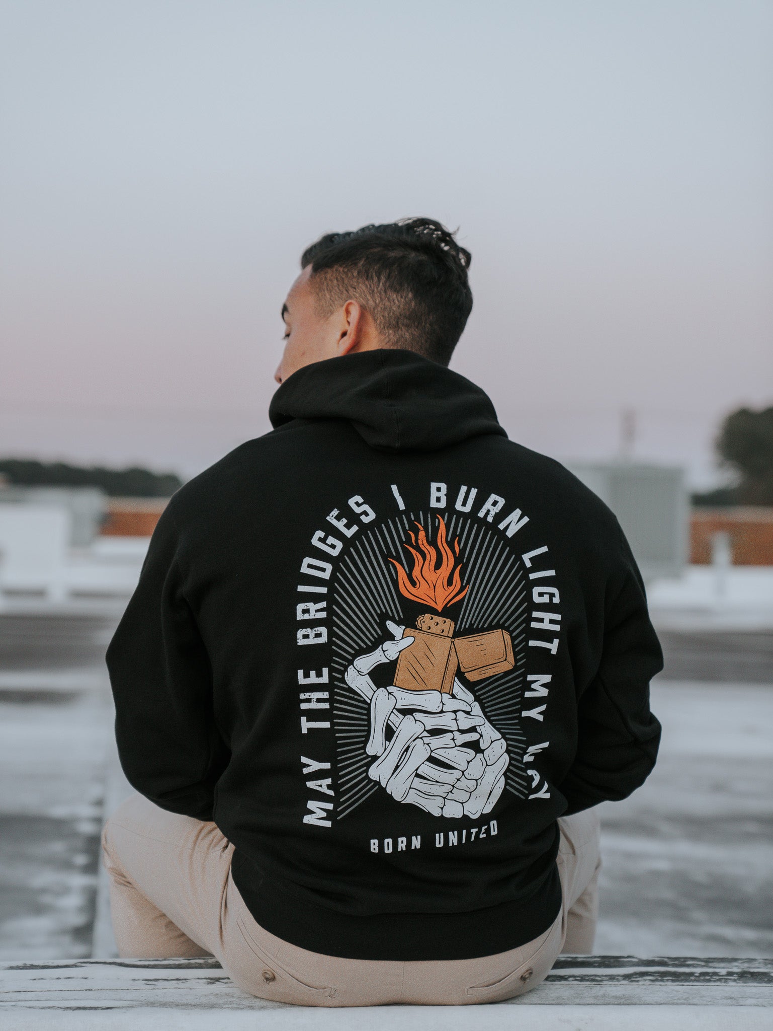A person with short hair sits outdoors, wearing the Burning Bridges Hoodie from Born United Private Label. The black hoodie features a graphic of hands holding a lit matchstick with the text: May the bridges I burn light my way, set against a soft, blurred sky.