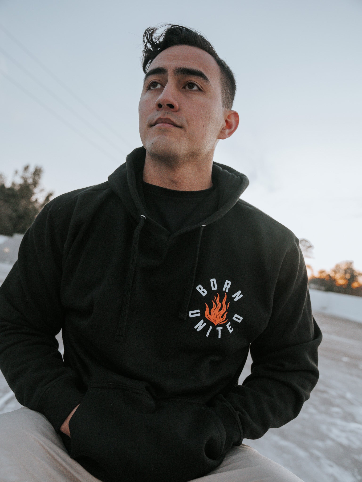 A person sits outdoors in daylight wearing a black Burning Bridges Hoodie from Born United Private Label, featuring Born United and a flame logo. With short dark hair, they gaze into the distance where trees stand silently in the background.