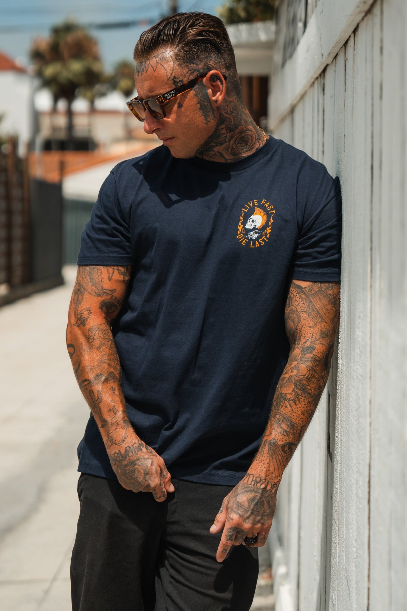 A tattooed man with sunglasses and slicked-back hair embodies youth culture as he leans against a white fence, wearing Relentless Betrayals Live Fast Navy T-Shirt and black pants, exuding the live fast, die last ethos amidst palm trees and blurred buildings.