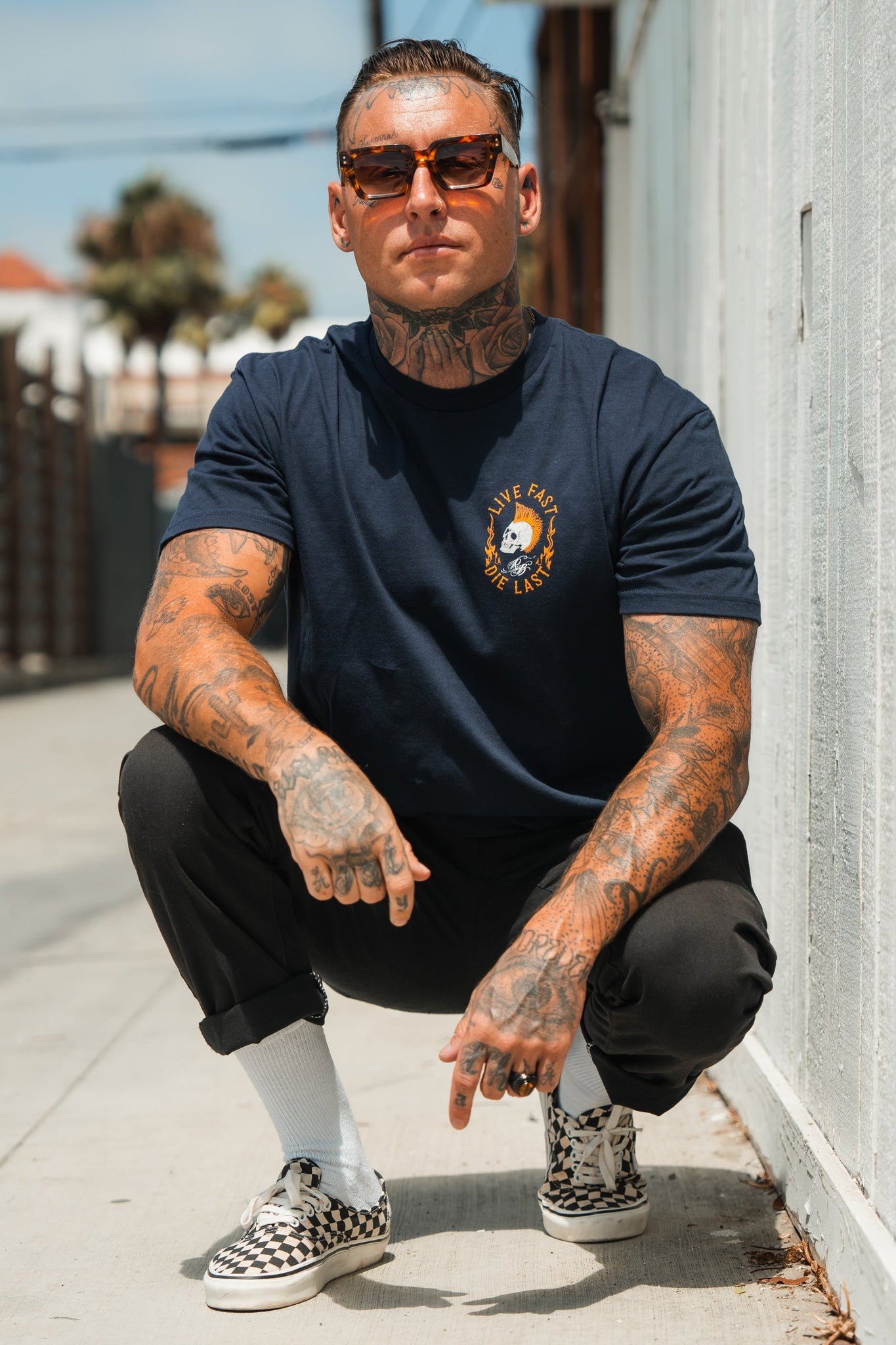 A tattooed man embraces youth culture, crouching on a sidewalk. Wearing sunglasses, Relentless Betrayals Live Fast navy T-shirt, black pants, white socks, and checkered sneakers, he embodies a carefree spirit with a fence and blurred trees creating an adventurous backdrop.