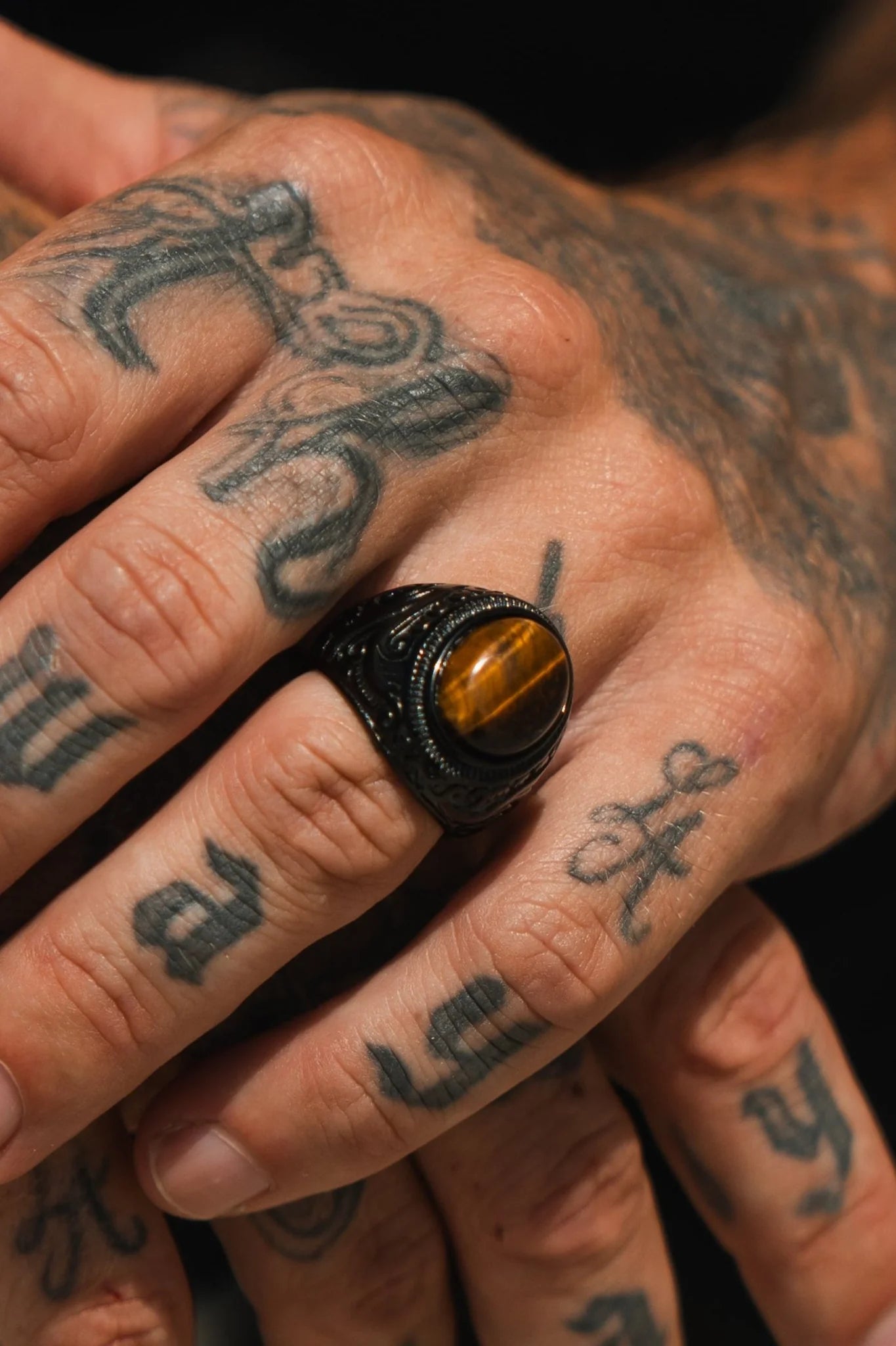 Tattooed hands display the Eye Of The Tiger Ring by Relentless Betrayal, featuring a large oval tigers eye stone set in stainless steel. The tattoos and a blurred background accentuate this unique piece of jewelry.