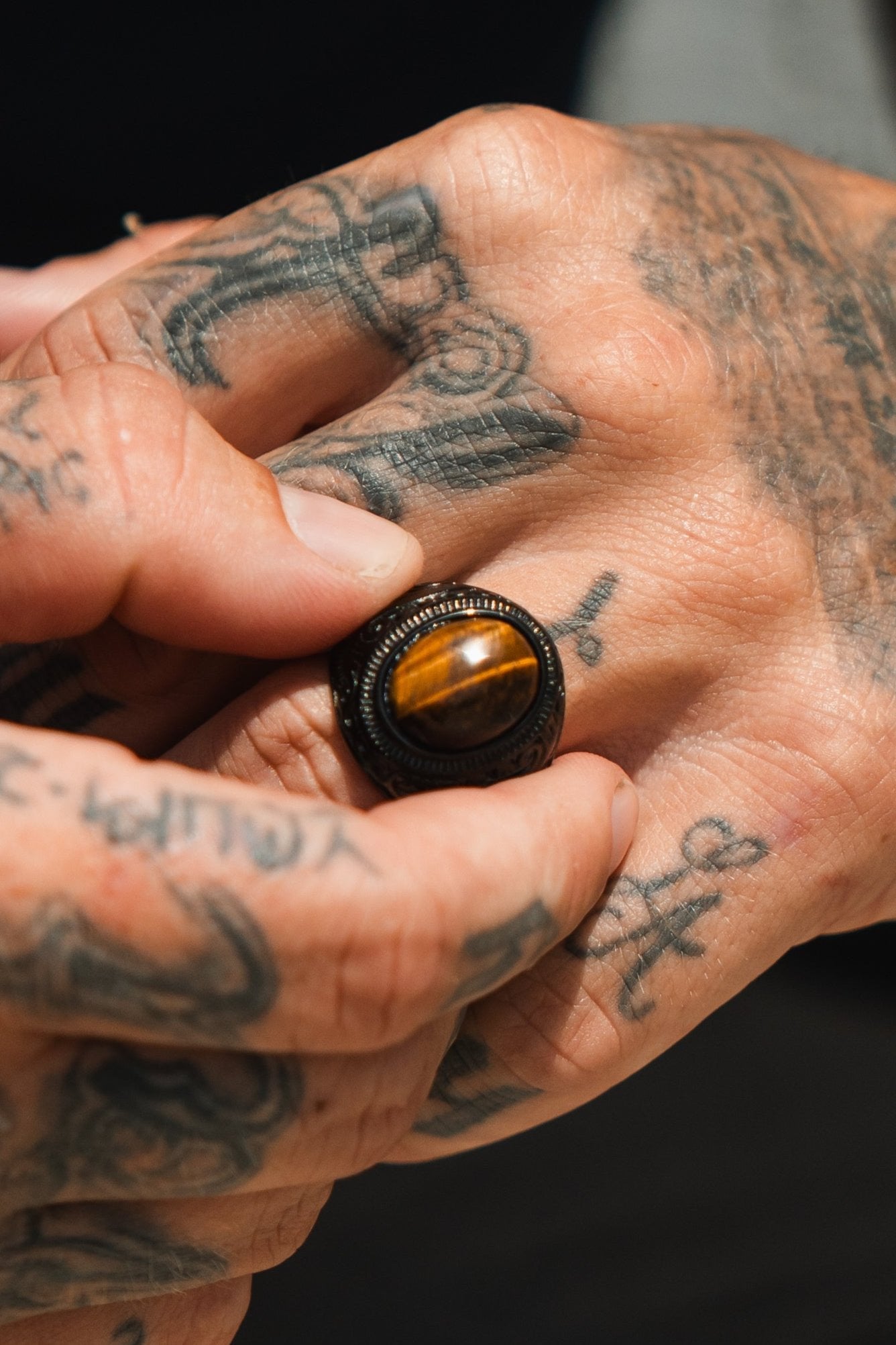 Tattooed hands expertly adjust the Eye Of The Tiger Ring by Relentless Betrayal, showcasing its ornate, dark metallic setting that evokes the look of stainless steel.