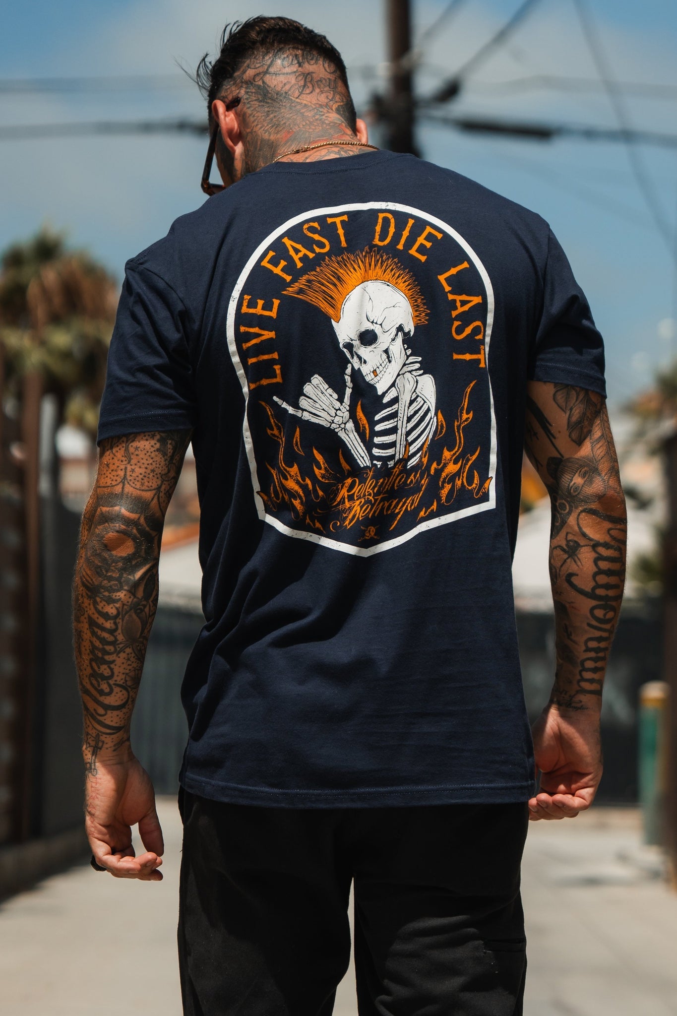 A tattooed individual embodies youth culture, facing away in a Relentless Betrayal Live Fast Navy T-Shirt featuring a skeleton and flames with the text Live Fast Die Last, standing ready for adventure under a clear outdoor sky.