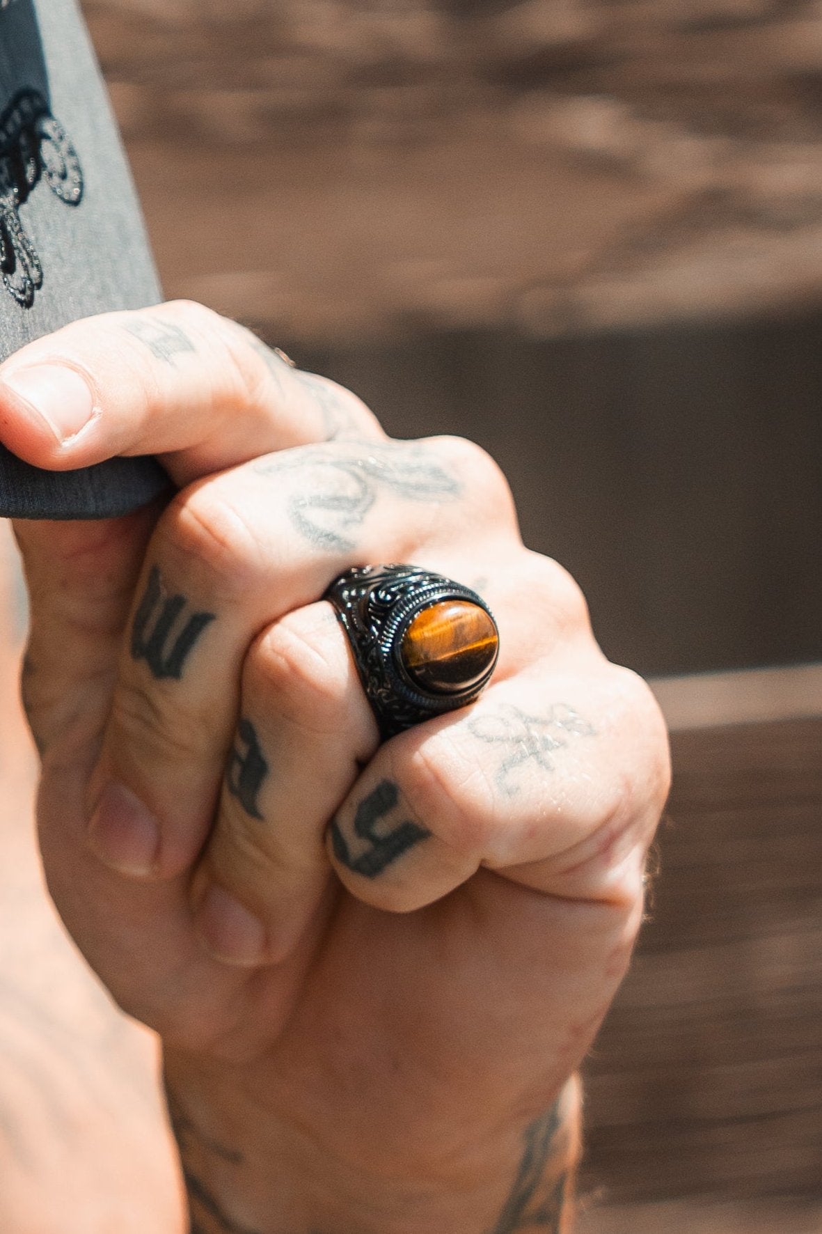 A tattooed hand grips a skateboard while wearing Relentless Betrayals Eye Of The Tiger Ring, a black stainless steel piece highlighted by a striking tiger eye gemstone. The blurred background shifts focus to the handcrafted jewelrys detailed design.
