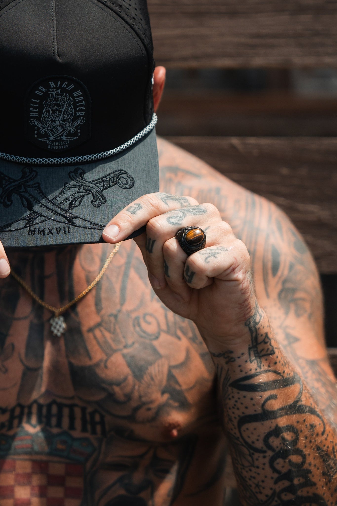 A tattooed person holds the brim of a cap, hiding their face. Theyre wearing the handcrafted Relentless Betrayals Eye Of The Tiger Ring and a gold chain necklace. The cap displays embroidered swords and text against wooden planks.